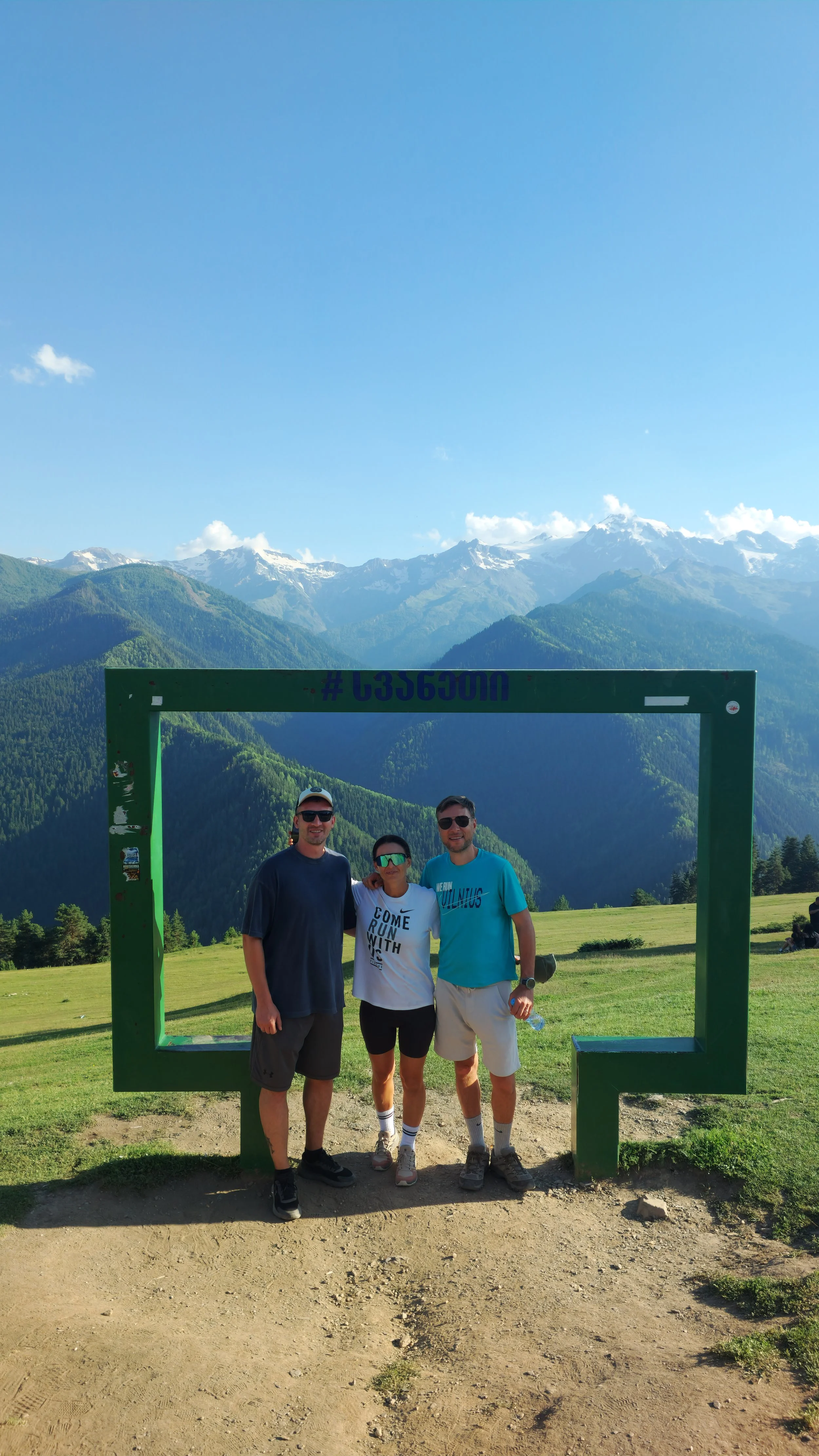 Three people standing inside a green square frame in a mountain landscape with green hills and snow-capped peaks in the background.