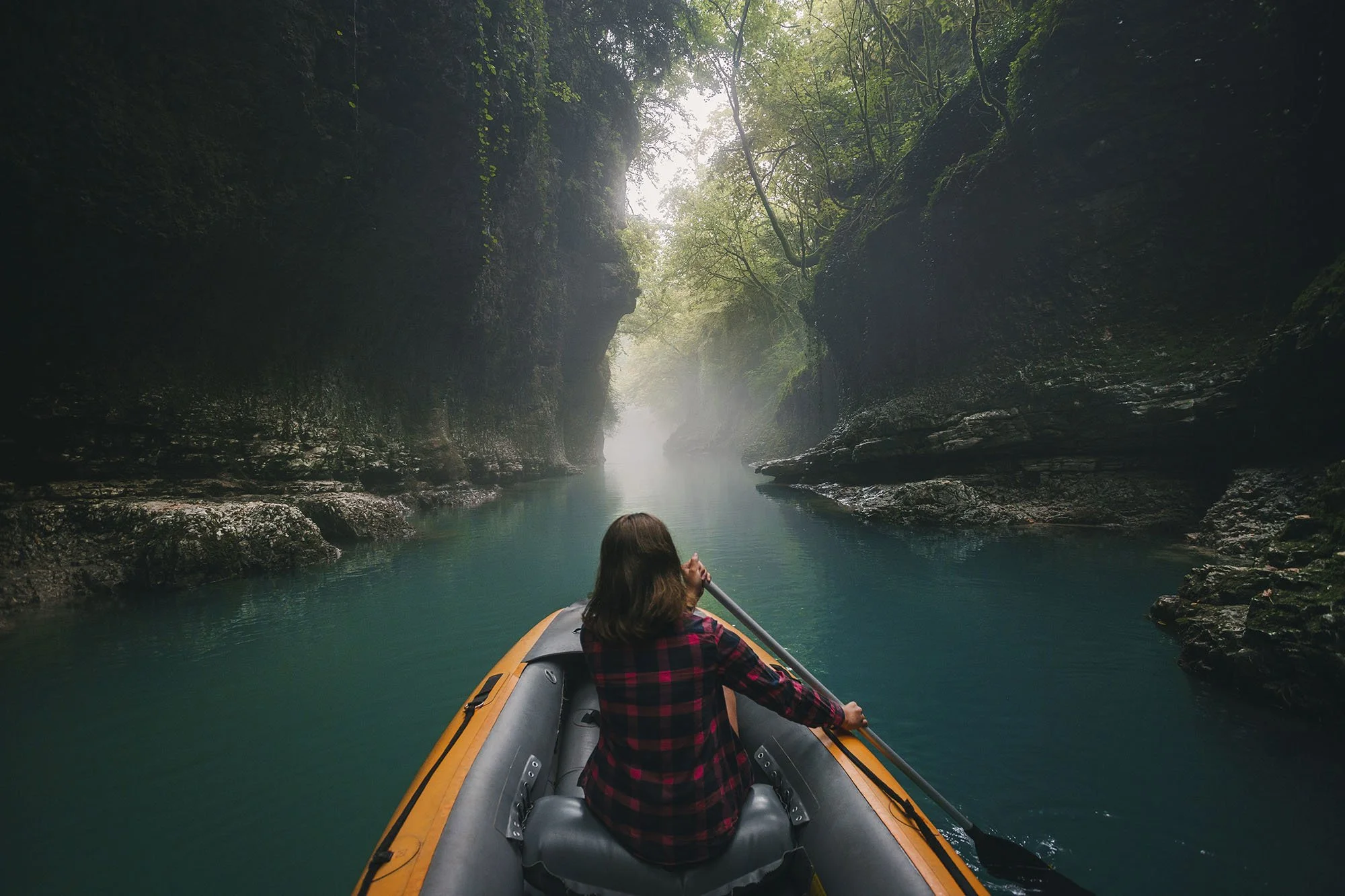 A woman kayaking through a narrow canyon with tall rocky walls and green foliage, overlooking a calm, blue river.