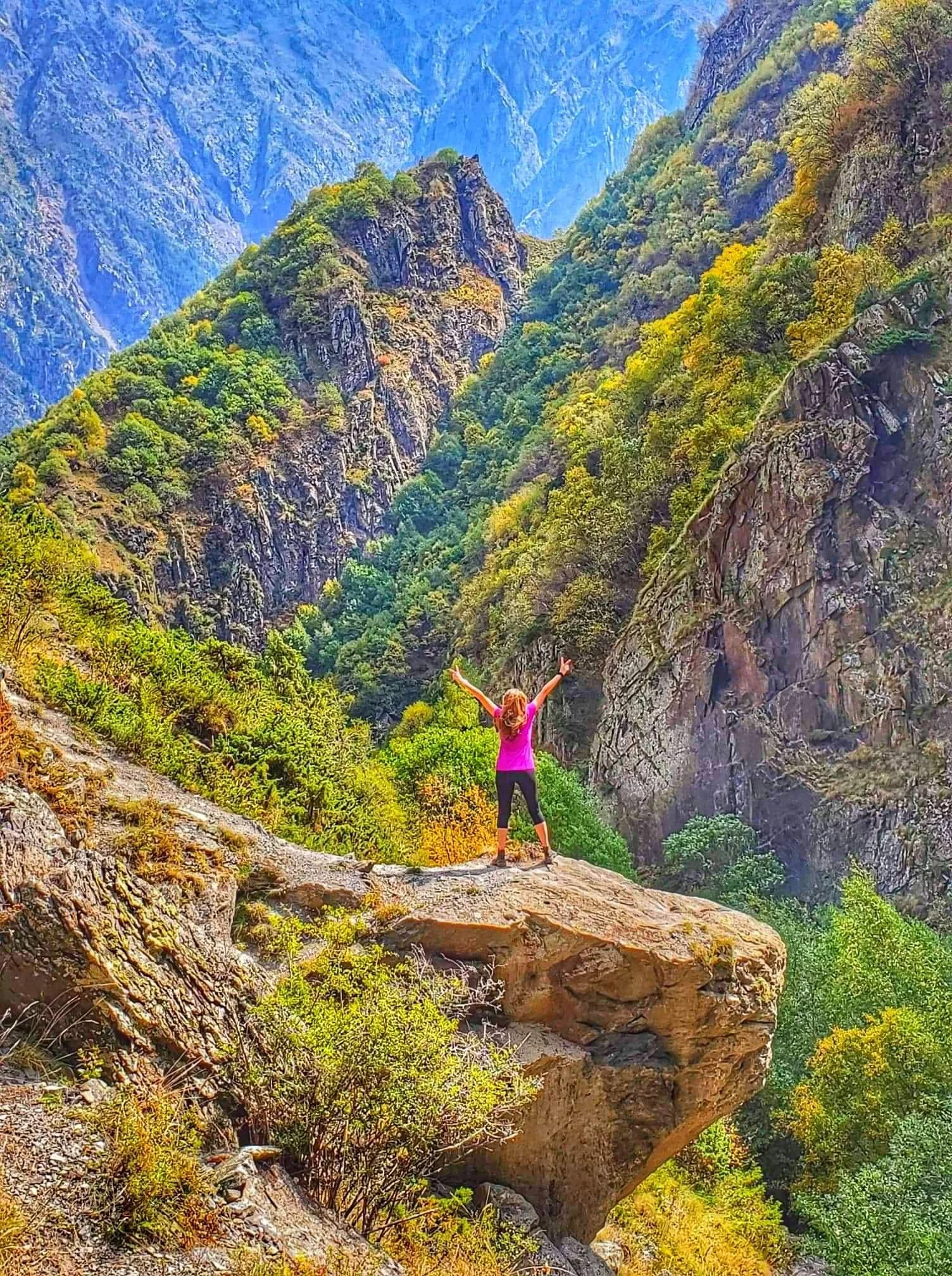 A woman in a pink shirt and black pants standing on a rock ledge with arms raised, overlooking a lush, green mountain landscape with tall cliffs and valley.