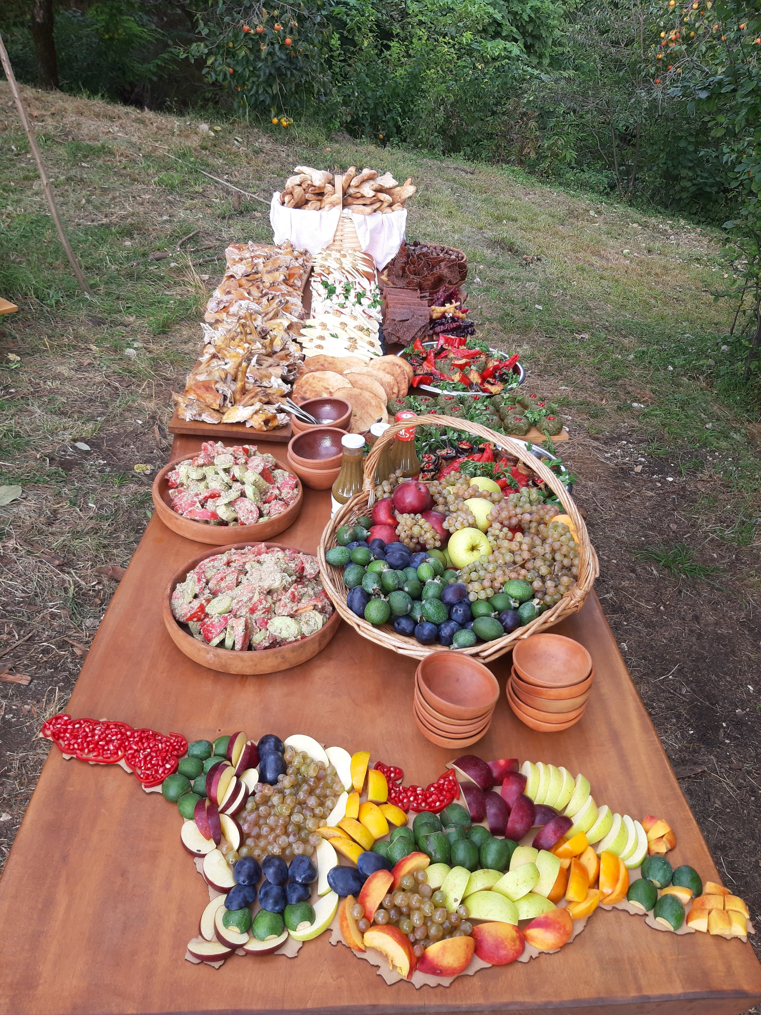 A long outdoor table displays an assortment of fresh fruits, baked goods, and other foods, with trees and greenery in the background.