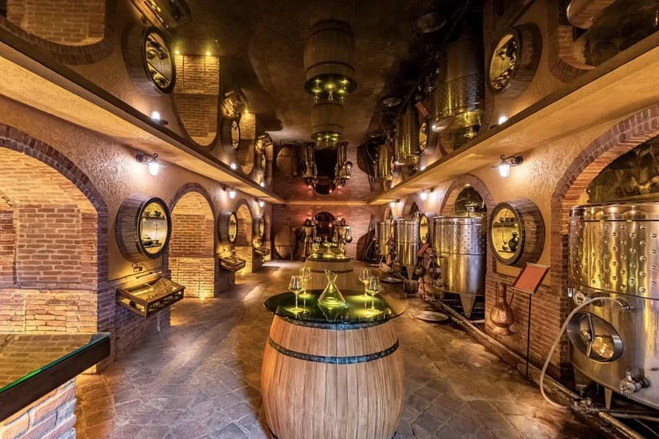 A winery or brewery with stainless steel tanks and equipment, brick walls, and a barrel table with wine glasses.