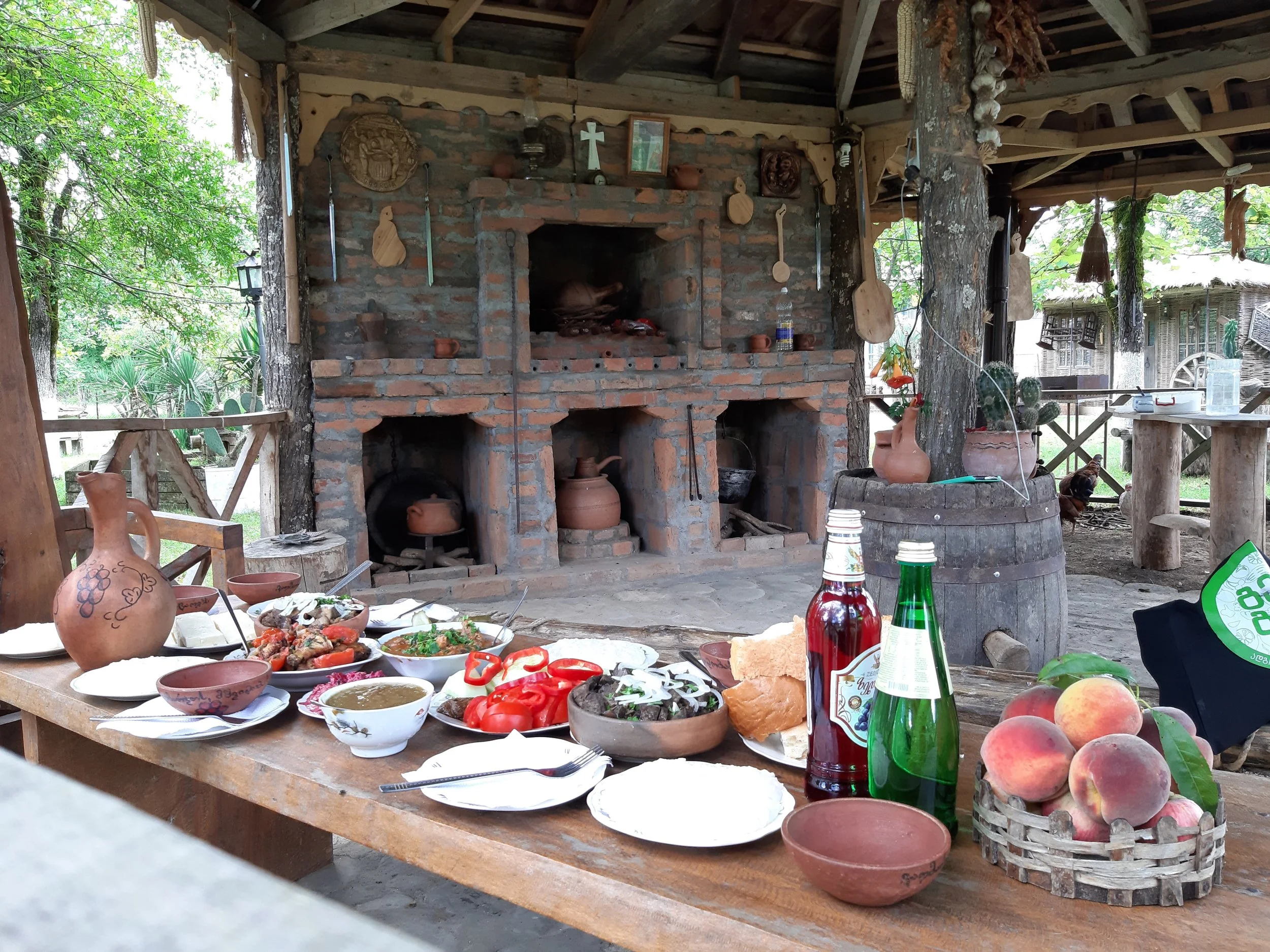 A rustic outdoor dining setup with a wooden table filled with various dishes, bowls, and beverages, set against a brick oven and brick wall with hanging decorative utensils, surrounded by trees and outdoor structures.