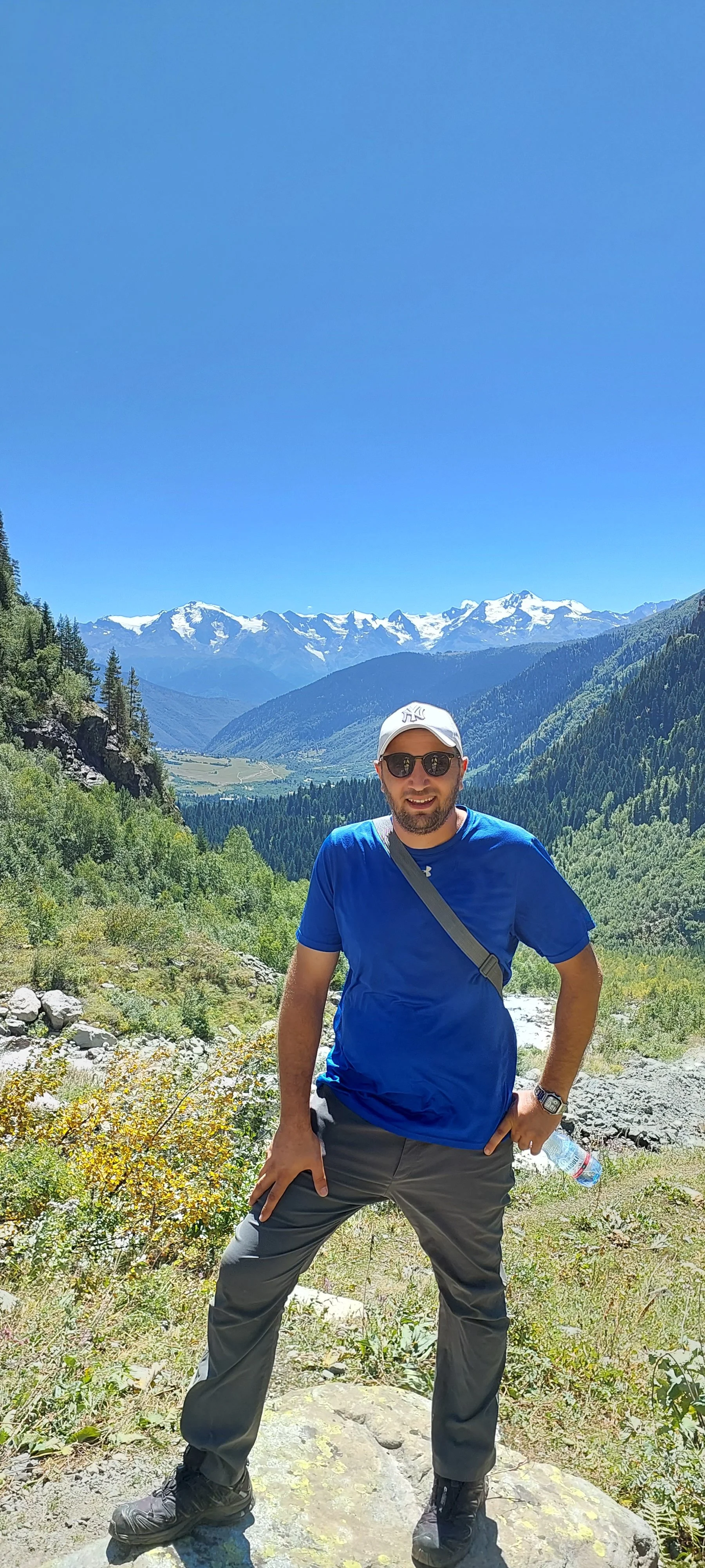 A man standing outdoors on a large rock, outdoors in a mountainous area with snow-capped peaks, green forested hills, and bright blue sky in the background.