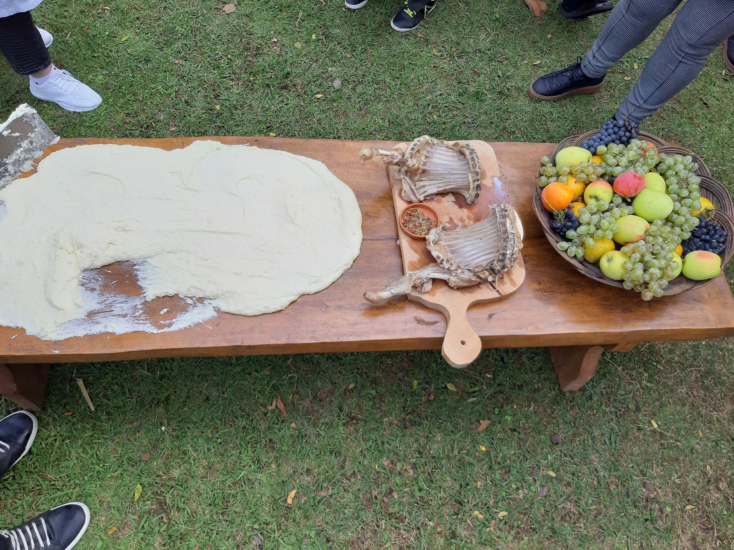 A wooden table with rolled-out dough, two scallop shells, a bowl of grapes and apples, and some shoes visible around it.
