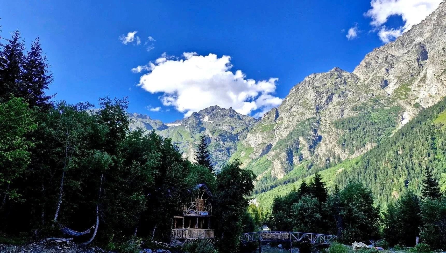 Scenic mountain landscape with lush green trees in the foreground, rugged mountains with patches of snow in the background, and a bright blue sky with scattered white clouds.