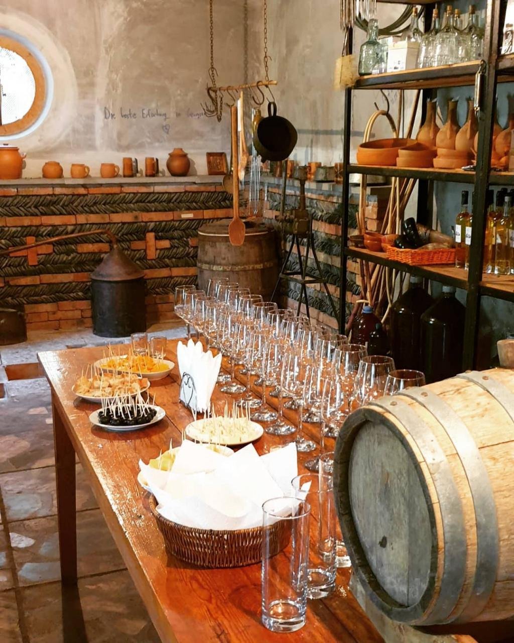 A rustic room set up for a wine tasting event with a long wooden table lined with empty wine glasses, plates of small appetizers with toothpicks, napkins, and a wine barrel, surrounded by shelves of pottery, glass bottles, and vintage kitchen items, with a chalkboard wall in the background.