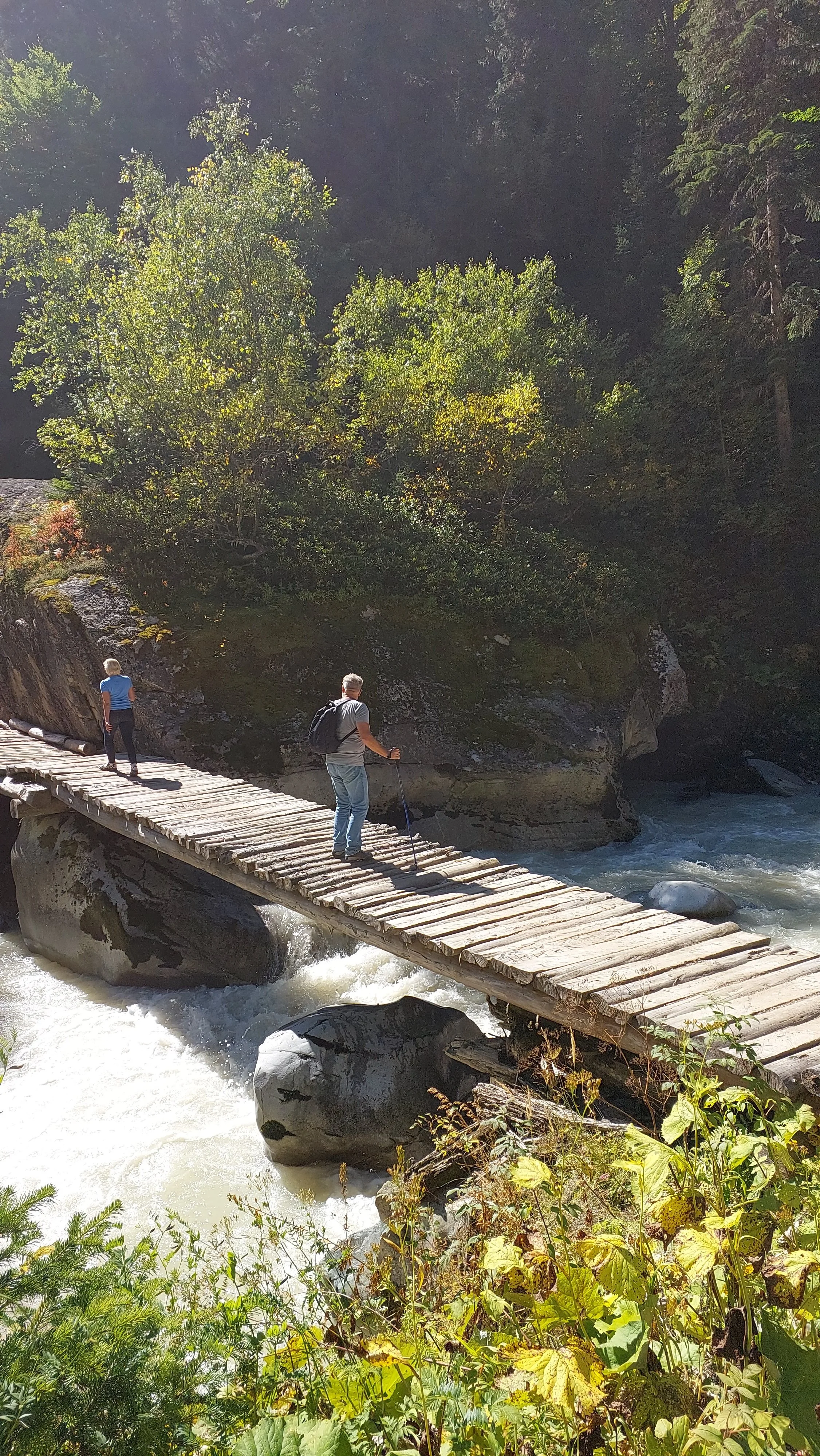 Two people crossing a wooden bridge over a rushing river in a forested area with trees and rocks.