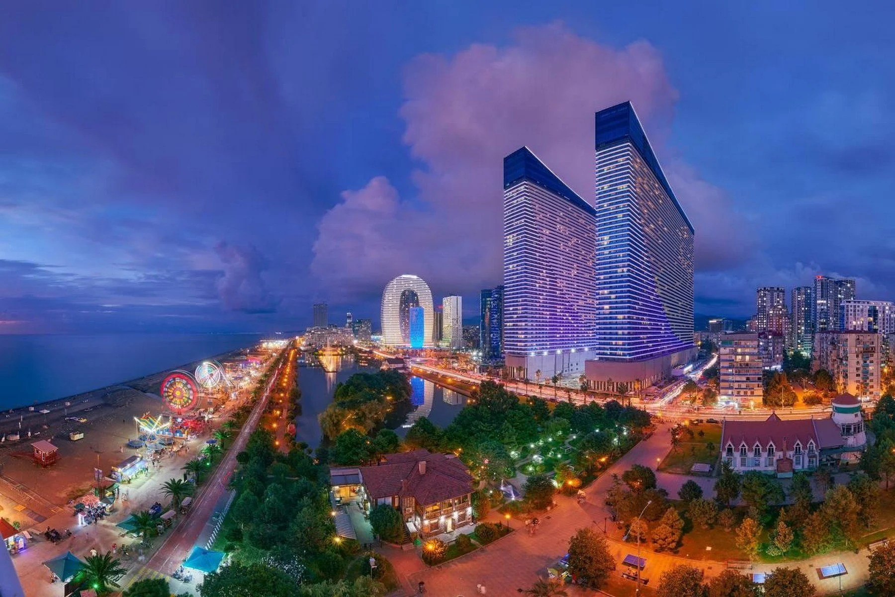 Nighttime cityscape featuring tall modern buildings, a waterfront, a park with trees and pathways, a small amusement area with lights, and a cloudy sky.