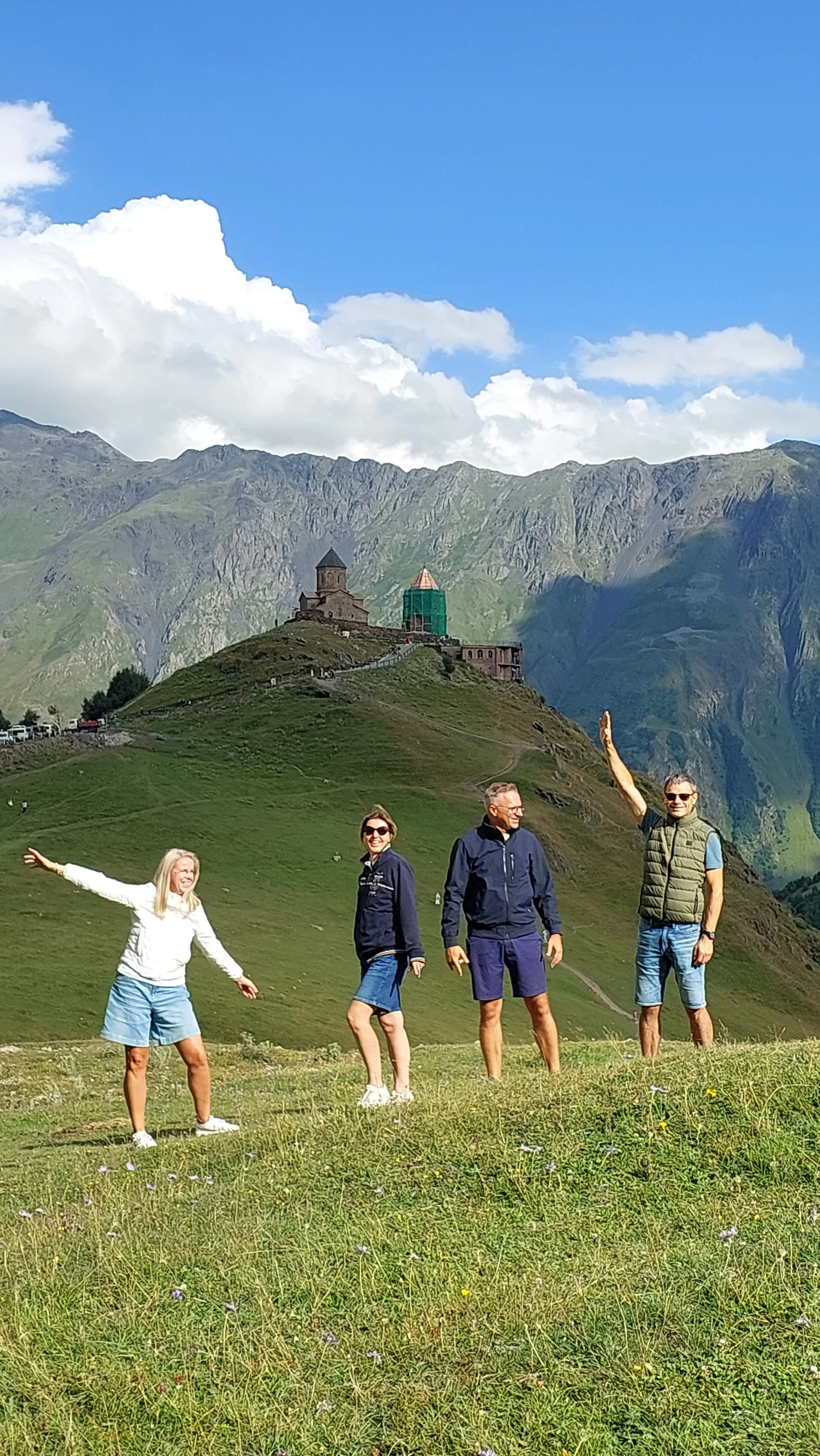 Four people standing on a grassy hill with a mountain and a historic church in the background under a partly cloudy blue sky.