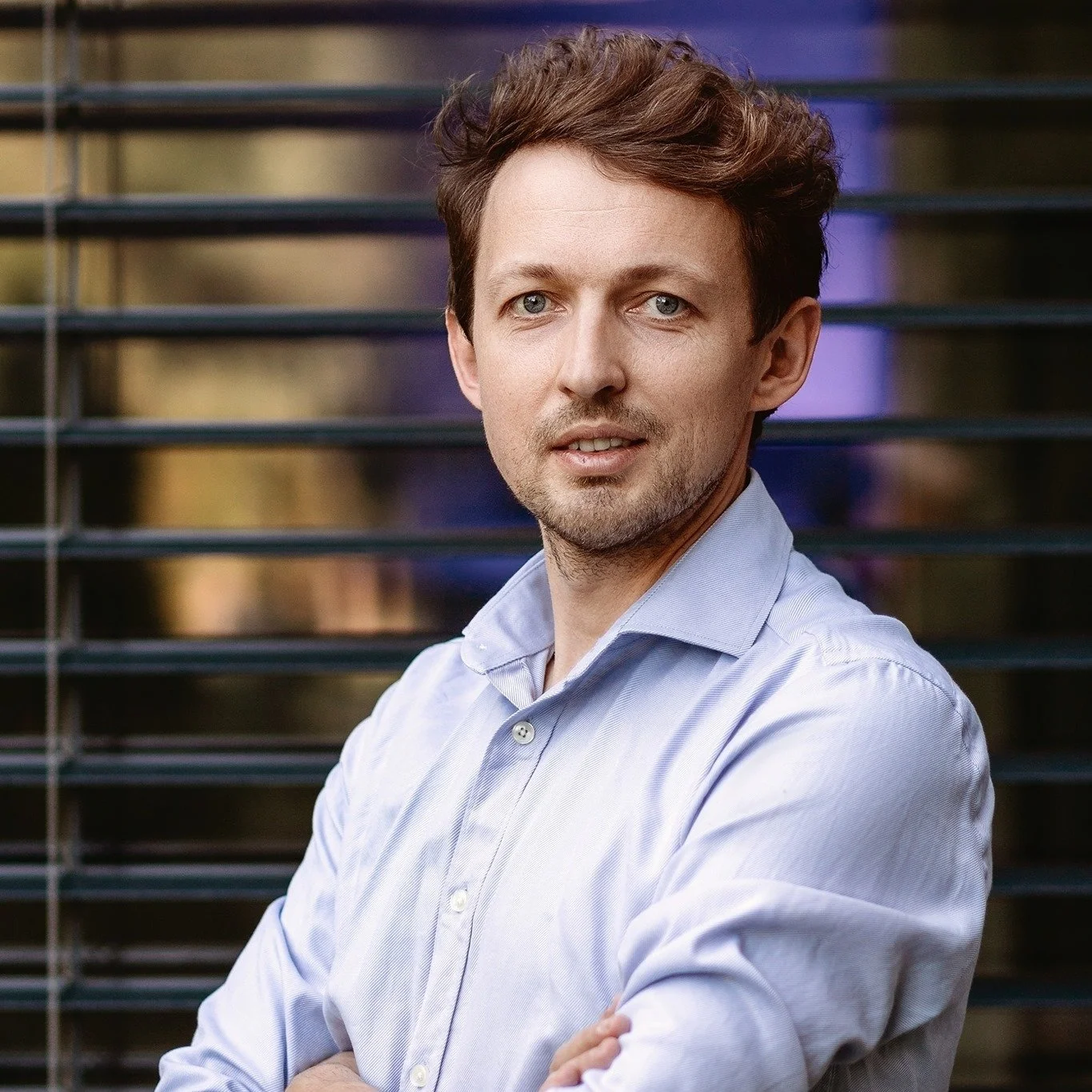 A man with light skin, brown wavy hair, and blue eyes wearing a light blue button-up shirt standing outdoors in front of a black metal railing with blurred city lights in the background.
