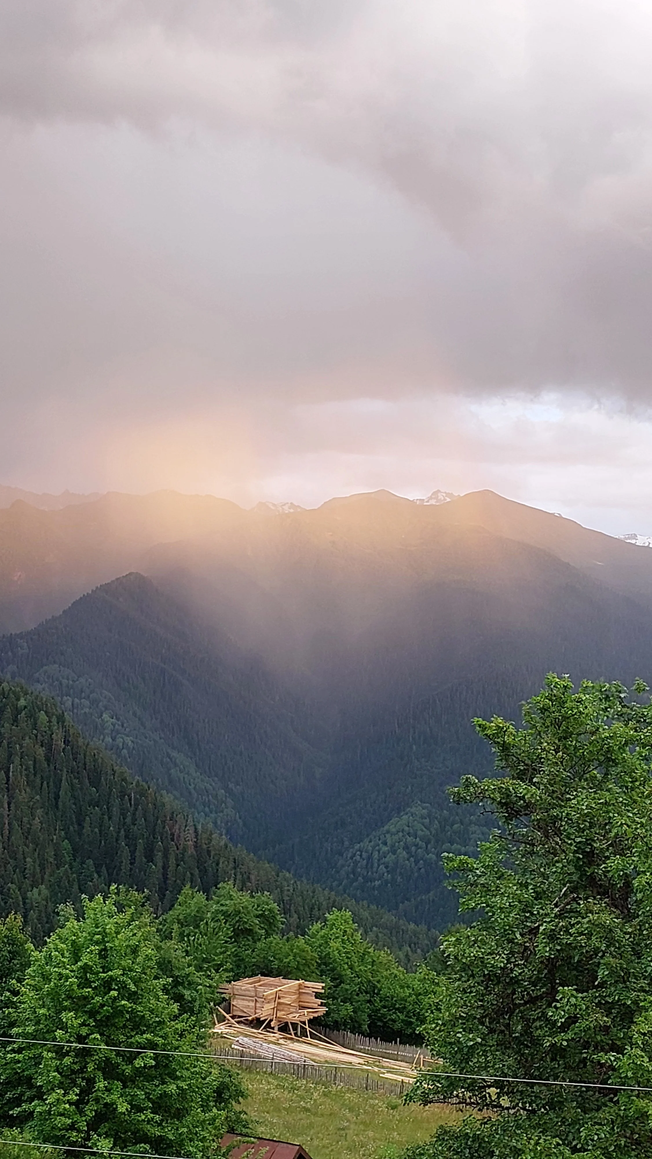 A scenic mountain landscape with green trees and a construction site with wooden planks in the foreground. The sky is cloudy with some sunlight breaking through.