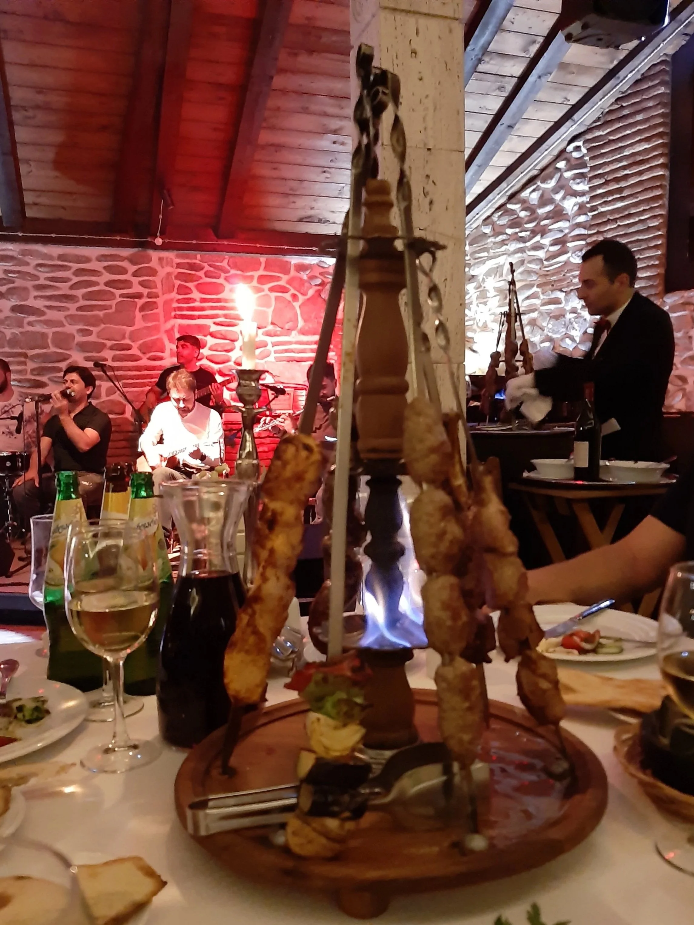 A dining table with grilled skewers, wine bottles, and glasses, with a live band performing in the background inside a rustic restaurant with stone walls and wooden ceiling.