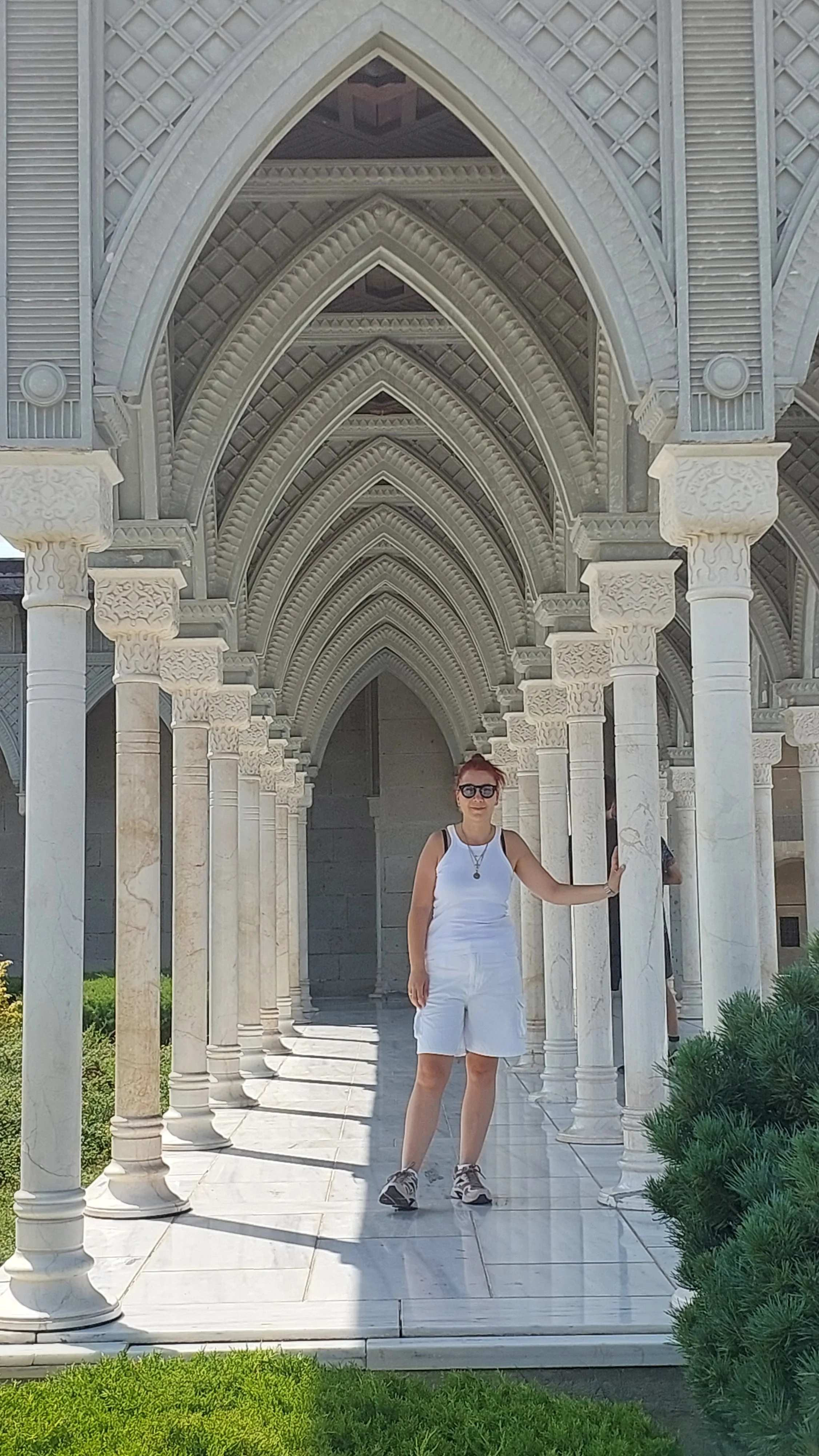 A woman in white shorts and tank top standing under a decorative stone archway with columns, holding onto one column, in a sunny outdoor setting.