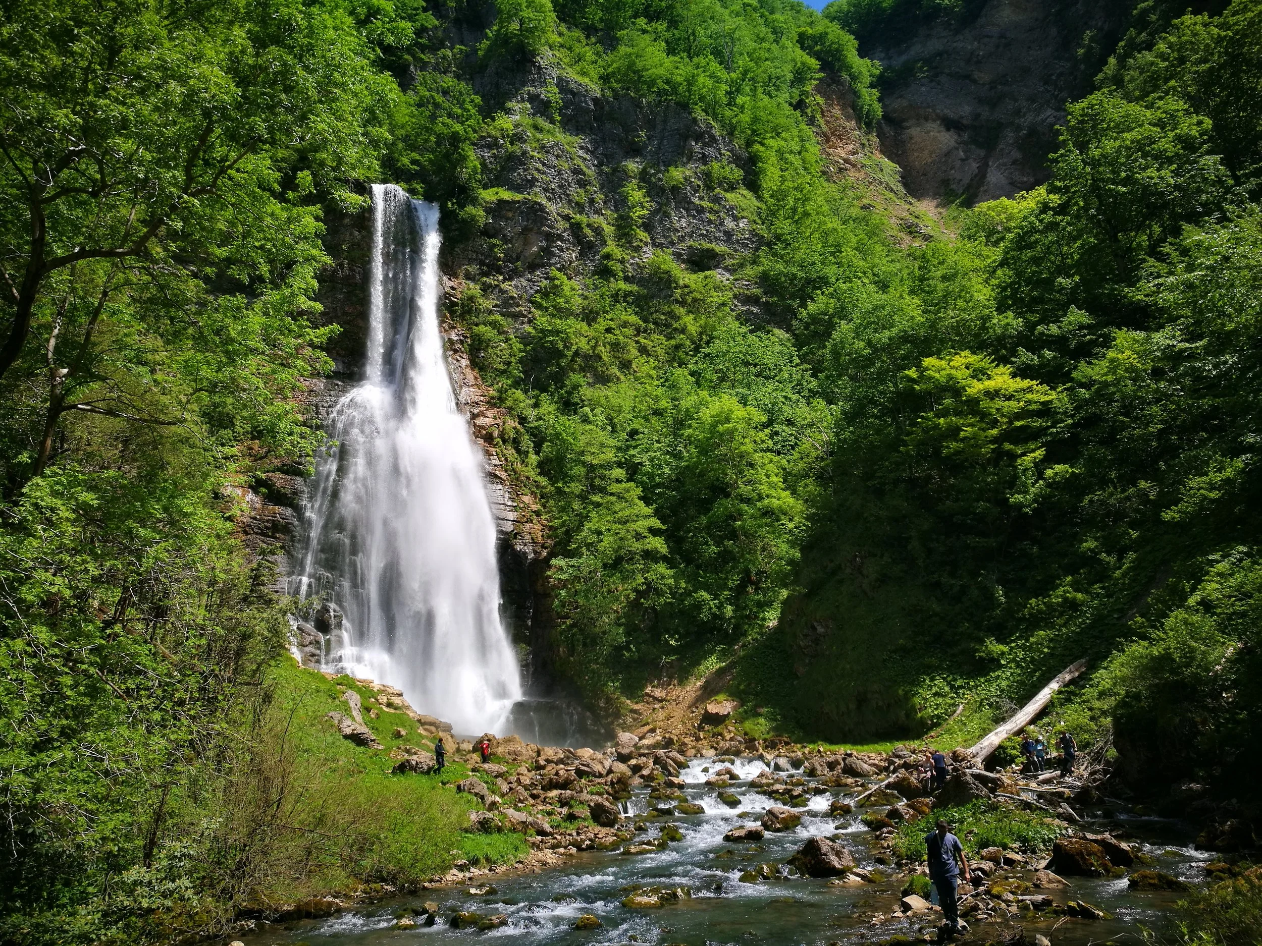 A tall waterfall cascading down a rocky cliff surrounded by green trees, with a stream flowing over rocks at the base and a few people exploring near the water.