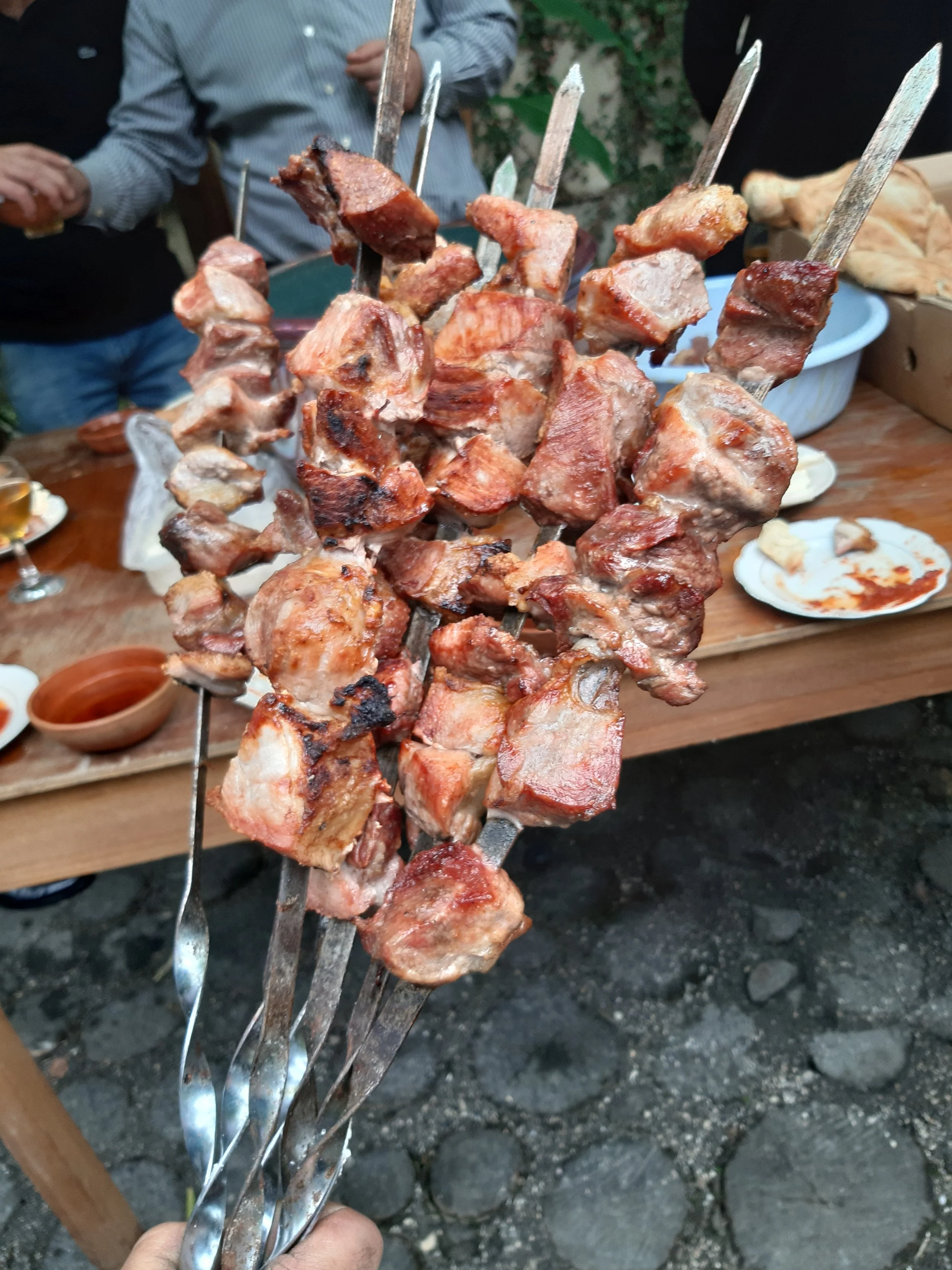 Close-up of skewers with grilled meat held up in front of people gathered around a table with plates and bowls of food outdoors.