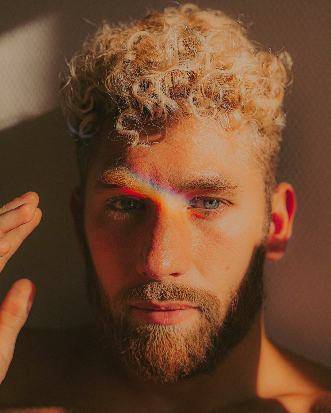 Un portrait d'un homme aux cheveux blonds bouclés et à la barbe, regardant fixement vers l'objectif, avec une lumière douce qui met en valeur ses traits.