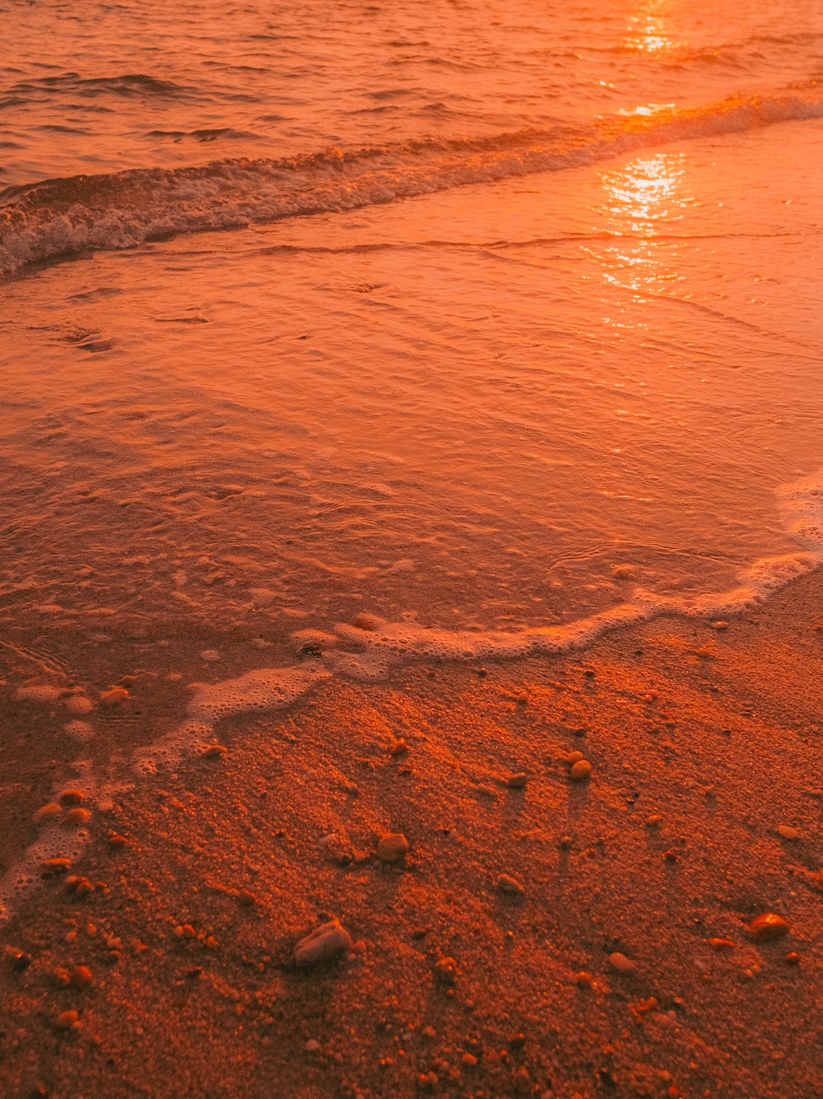 Plage de sable avec petites pierres sous un ciel rouge orangé lors du coucher ou lever du soleil, avec la mer calme et des vagues douces.