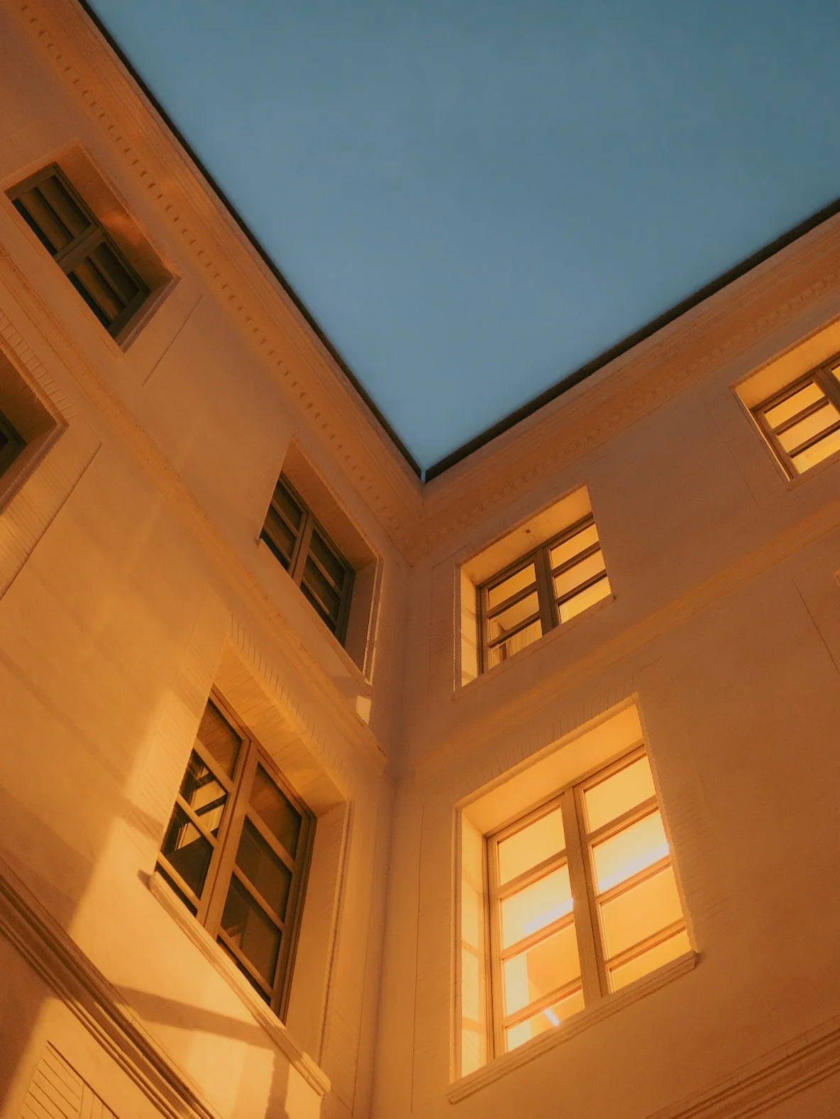 Vue intérieure d'un bâtiment vers le ciel, montrant une cour avec des fenêtres éclairées et un plafond ouvert, éclairé par une lumière chaude.