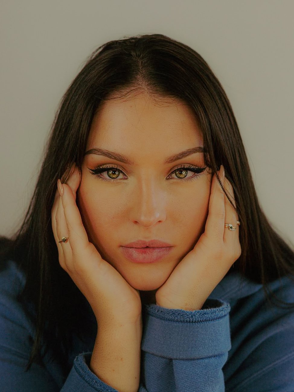 Une jeune femme aux cheveux noirs coiffés en ligne droite, portant un pull bleu, regarde directement l'appareil photo avec une expression sérieuse, ses mains soutenant son visage.