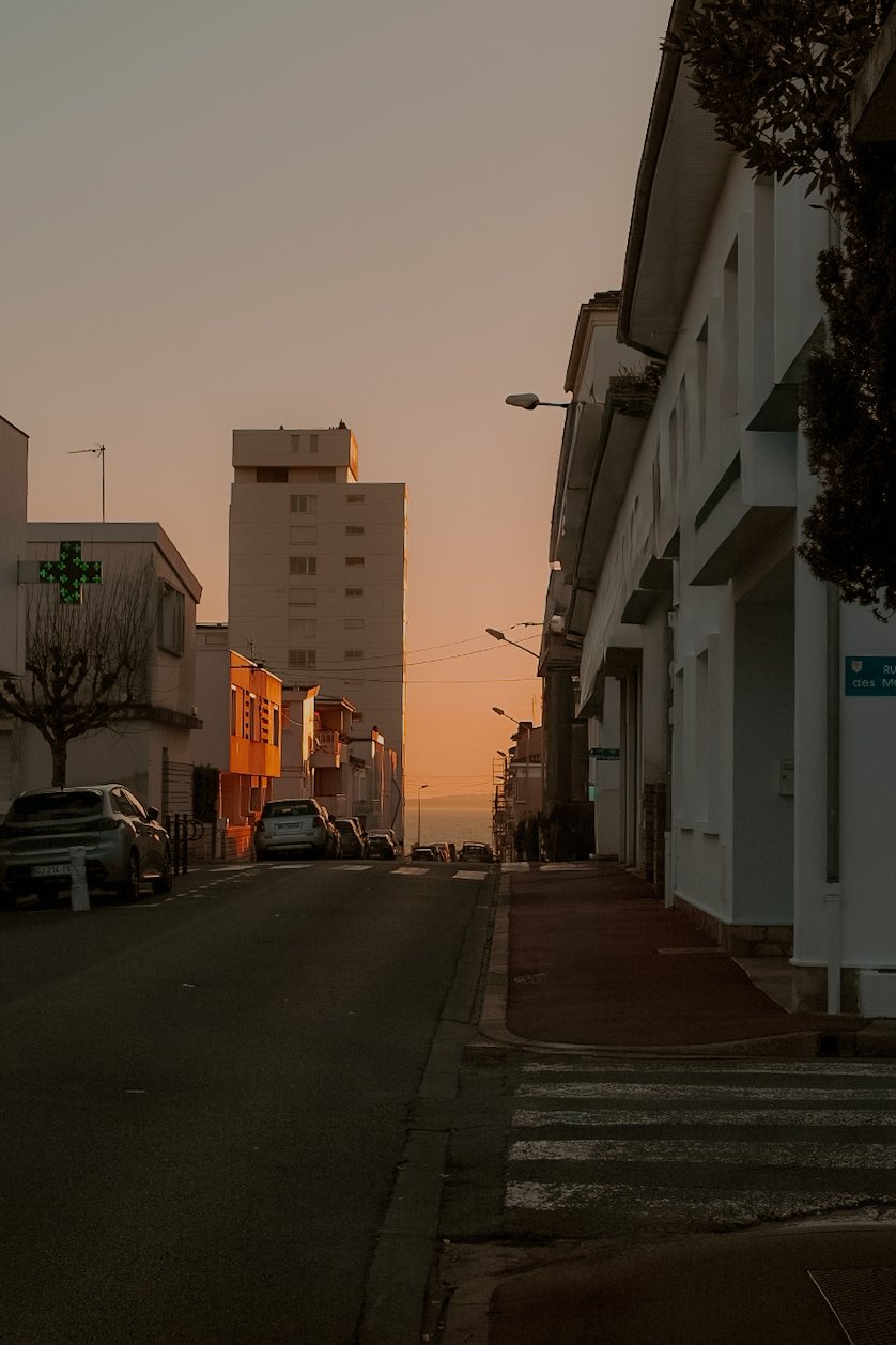 Une rue urbaine au coucher du soleil avec des bâtiments résidentiels et une pharmacie, vue vers l'océan au fond.