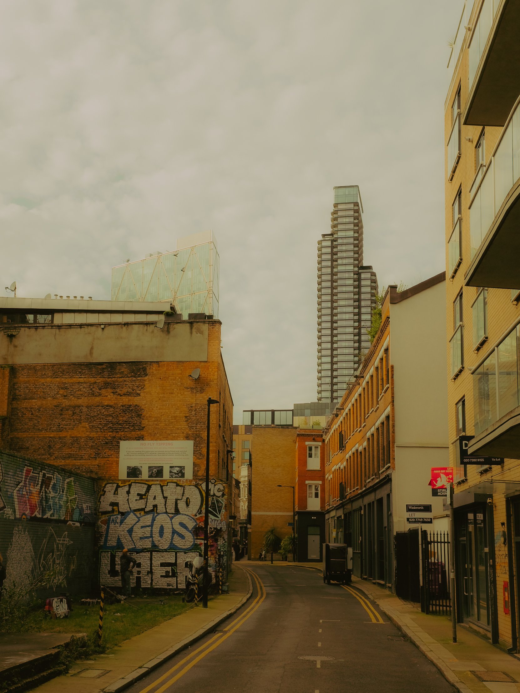 Rue urbaine bordée de bâtiments modernes et anciens, avec graffitis sur un mur à gauche, et gratte-ciel en arrière-plan sous un ciel nuageux.