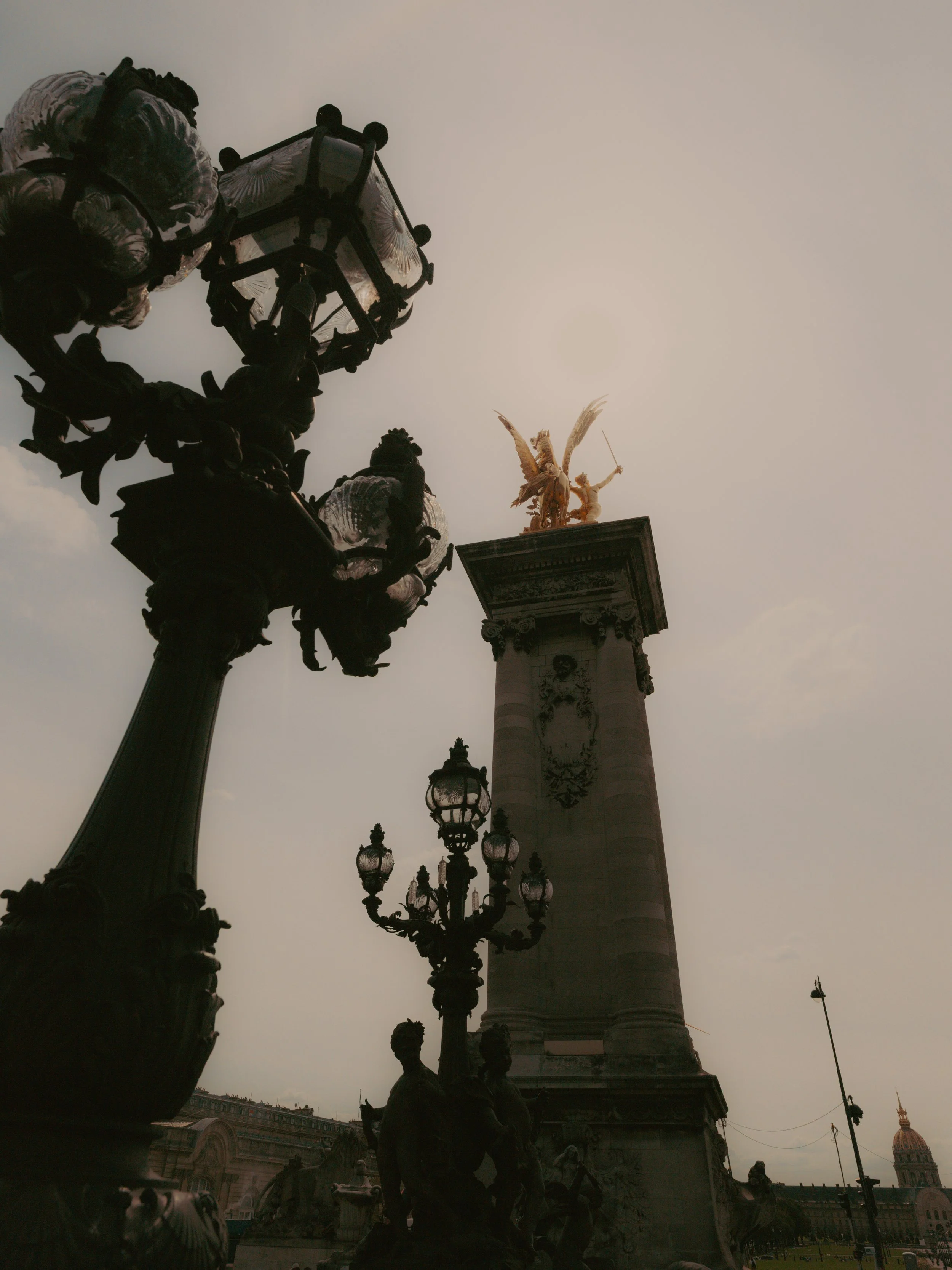 Une place publique avec une colonne ornée d'une sculpture dorée d'un ange avec des ailes déployées, entourée de lampadaires en fer forgé et de statues de figures humaines, avec un fond de bâtiments historiques.