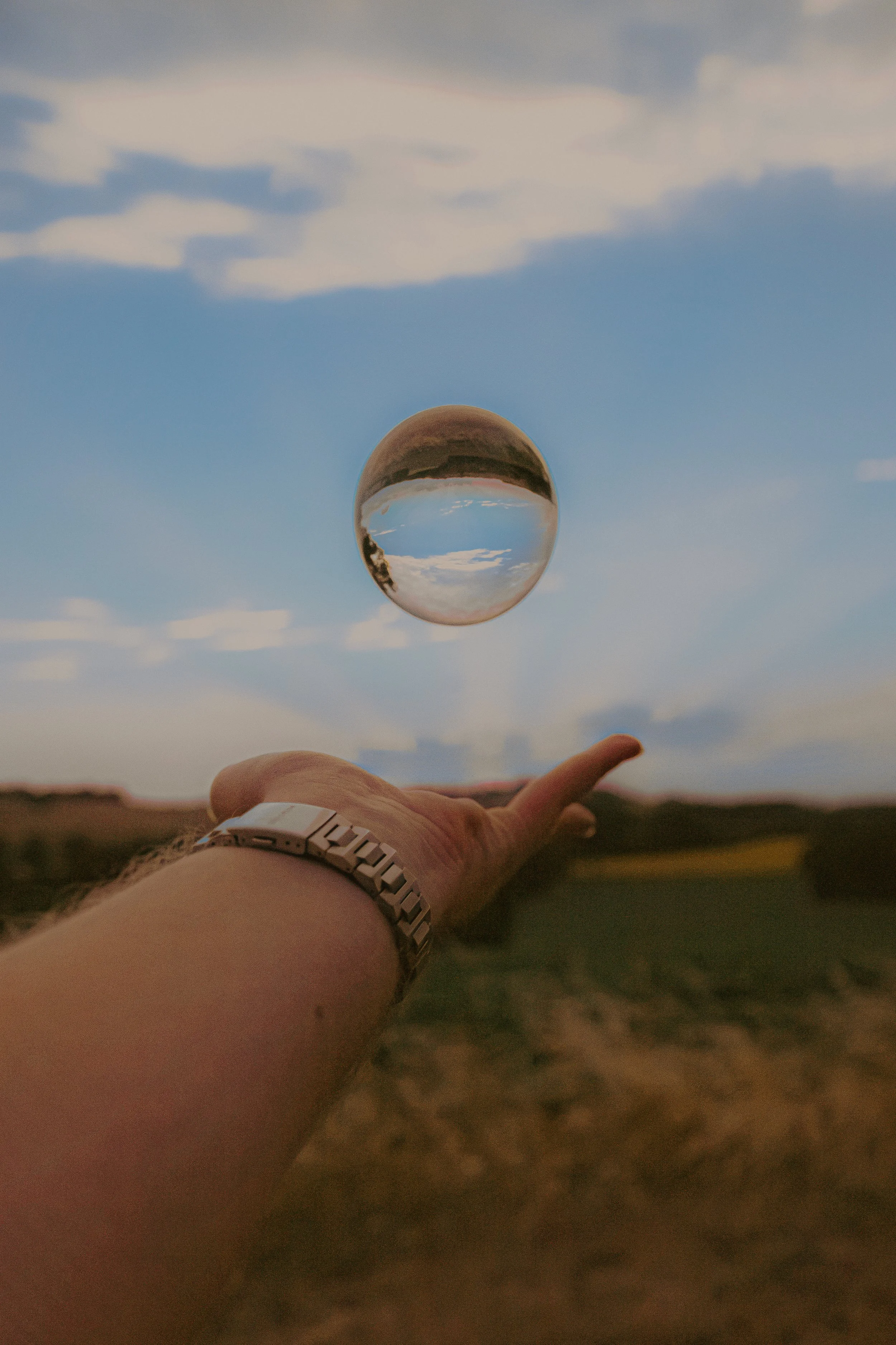 Une main portant une montre en acier pointe vers un ciel avec une boule en verre ou cristal suspendue, reflétant le ciel et le paysage environnant.