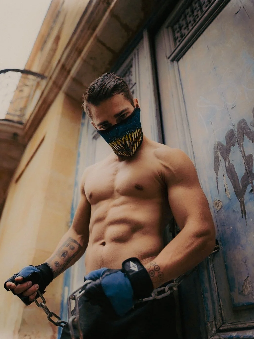 Un homme nu portant un masque facial, des gants de main et une chaîne enchaînée, se tenant dans une pièce avec des murs décorés et des fenêtres. 