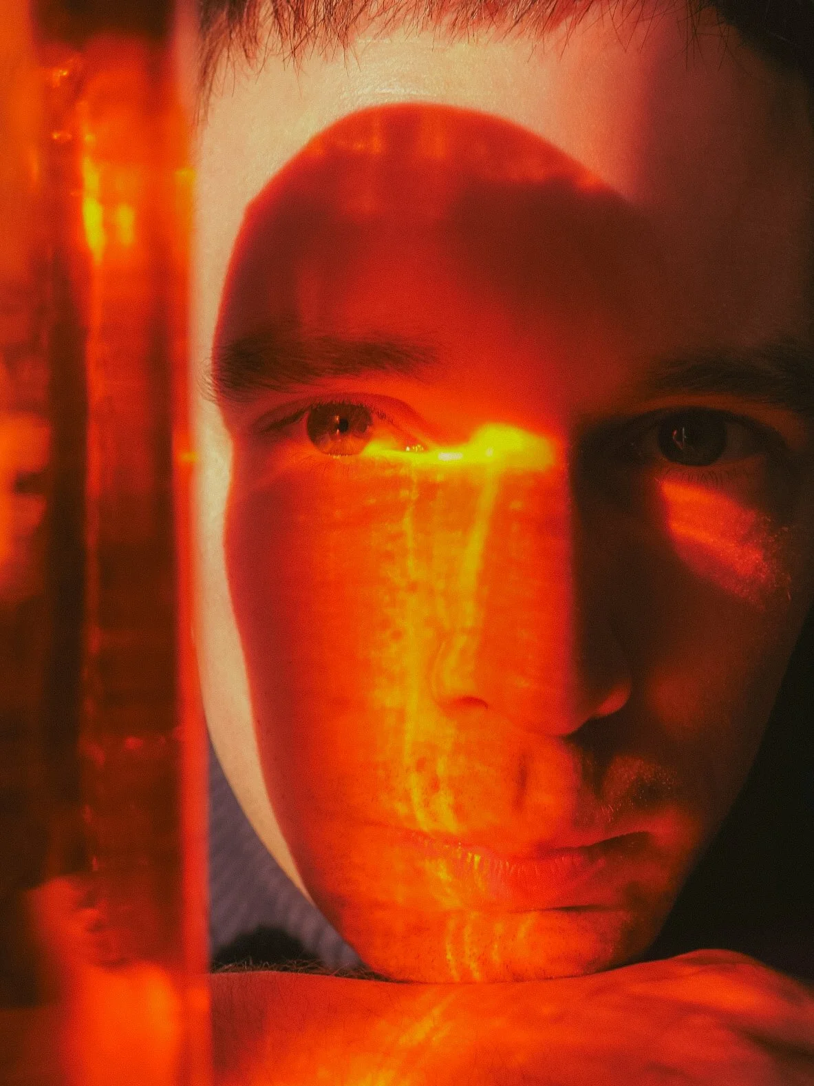 Premier plan d'un visage de jeune homme avec une expression sérieuse, illuminé par une lumière orange ou rouge, avec un effet de projection ou de reflet sur le visage.