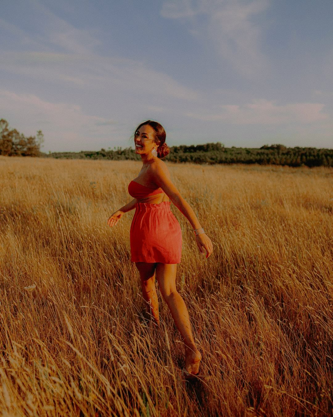 Une femme souriante marche dans un champ de blé doré au coucher du soleil, portant une robe rouge sans manches.