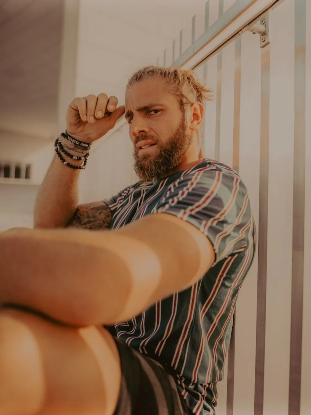 Un homme avec une barbe, portant un t-shirt à rayures, pose pour la photo en tenant sa main près de sa tête dans un endroit intérieur avec une rampes en métal derrière lui.