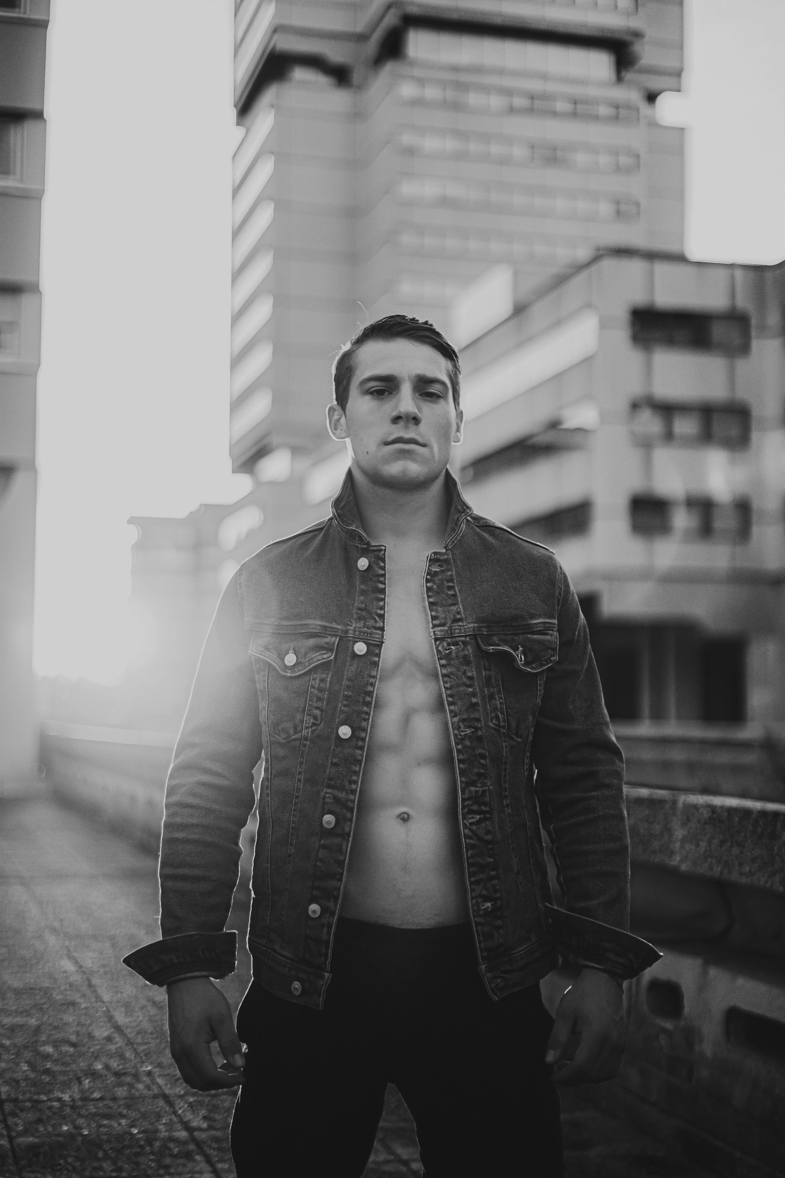 Un jeune homme debout dans une ville, portant une veste en jean ouverte, montrant son torse nu, avec un fond de bâtiments modernes en béton, en noir et blanc.