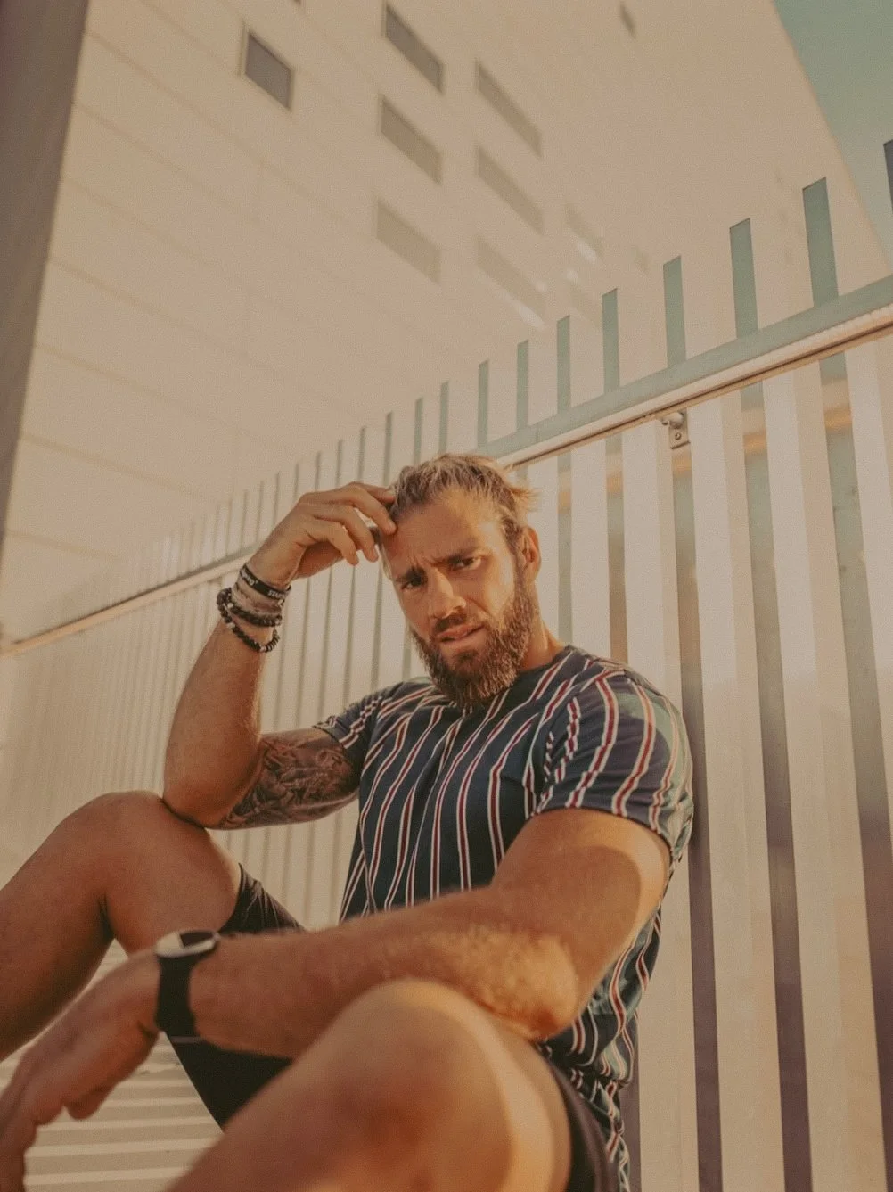 Un homme aux cheveux longs et barbe, portant un t-shirt à rayures, est assis devant une barrière en métal dans un environnement urbain, avec un bâtiment en arrière-plan.