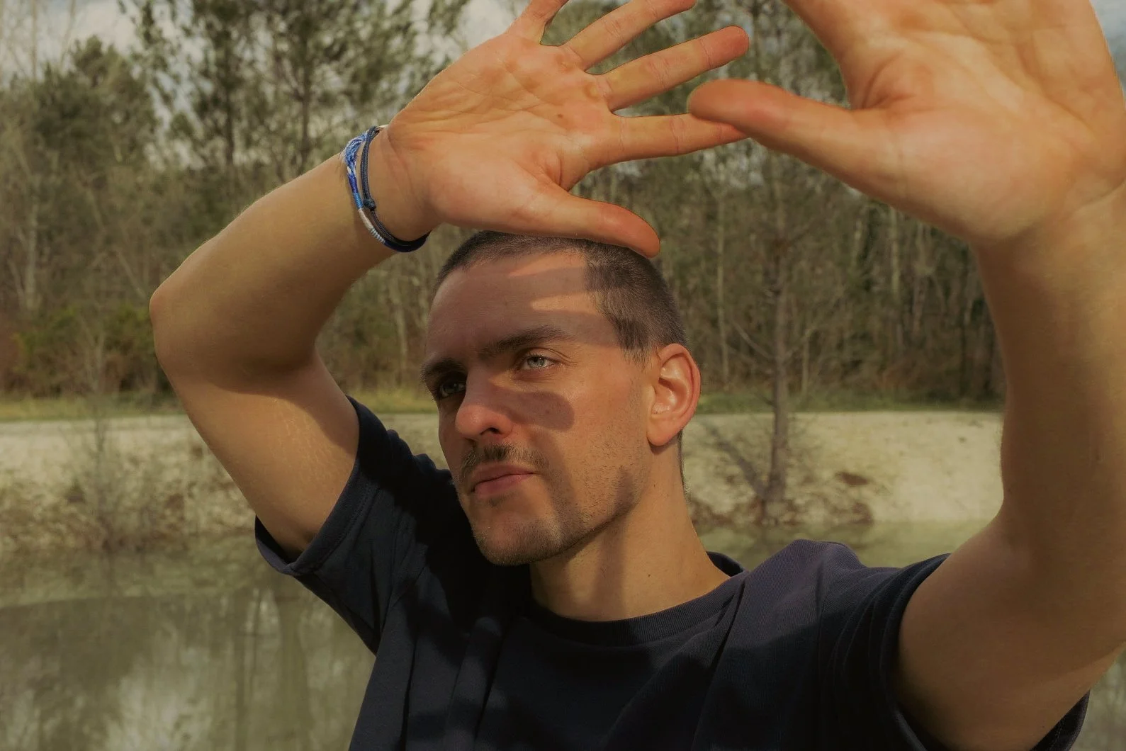 Homme avec regard pensif, mains levées, en extérieur près d'une rivière, arbres en arrière-plan.