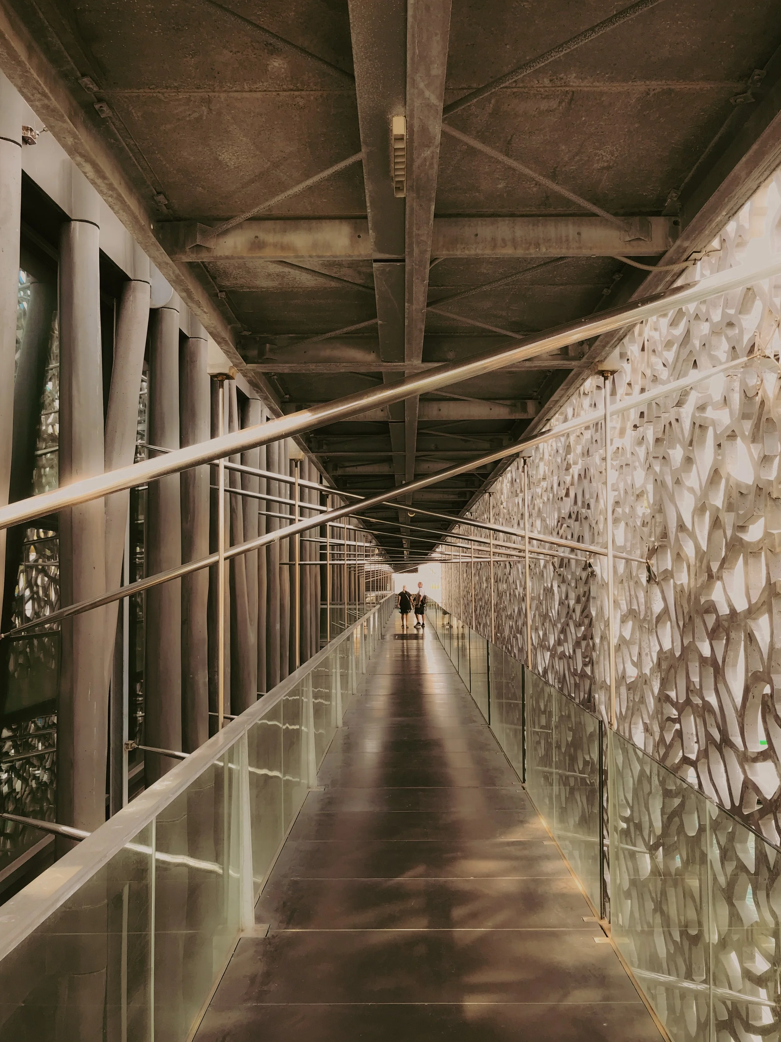 Passerelle en métal dans un bâtiment moderne, avec des murs décoratifs en relief coloré et des personnes en arrière-plan.