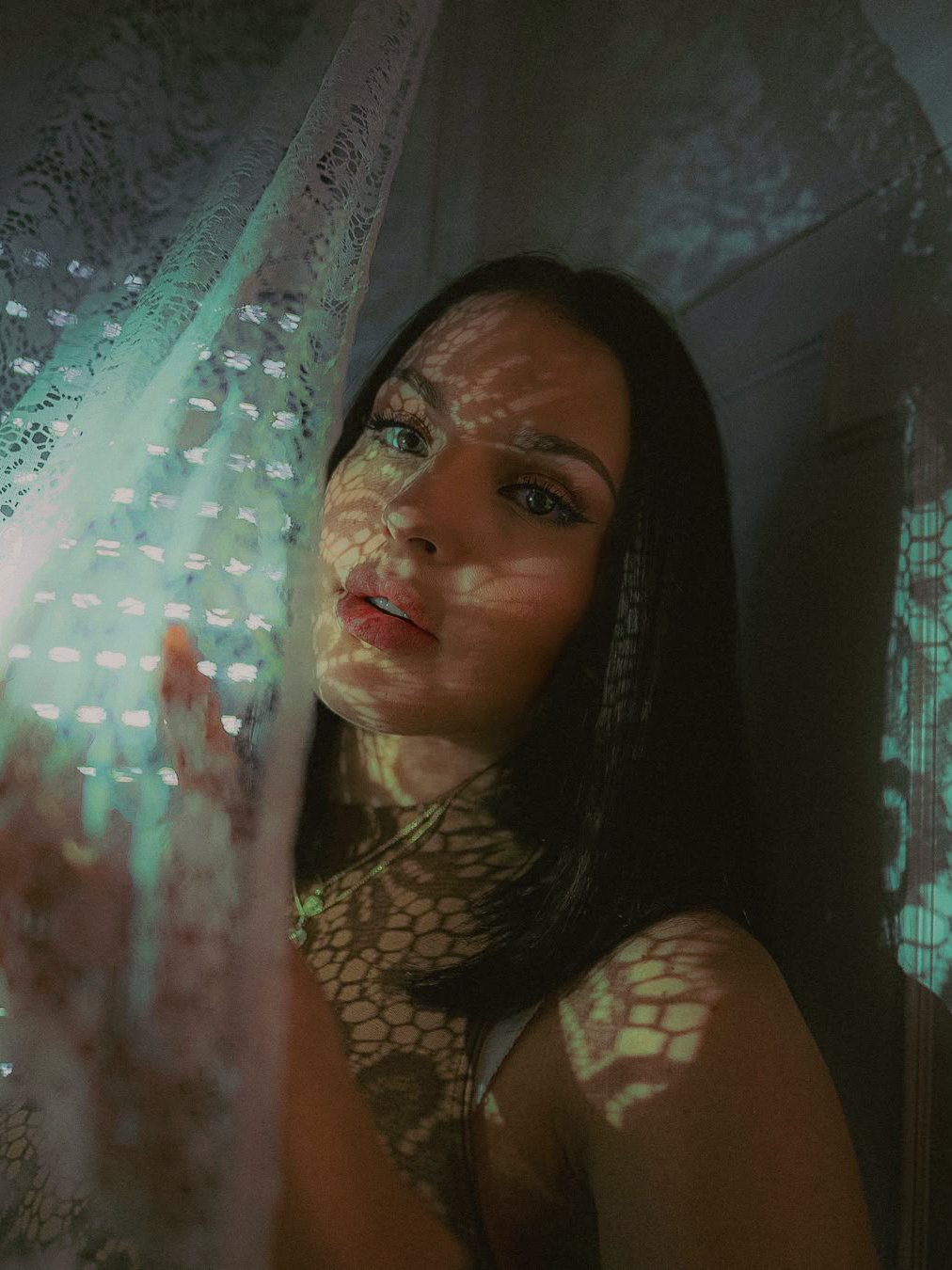 Une jeune femme aux cheveux noirs, regardant l'objectif, avec des motifs de lumières et d'ombres projetés sur elle et un drapé en dentelle à côté d'elle.