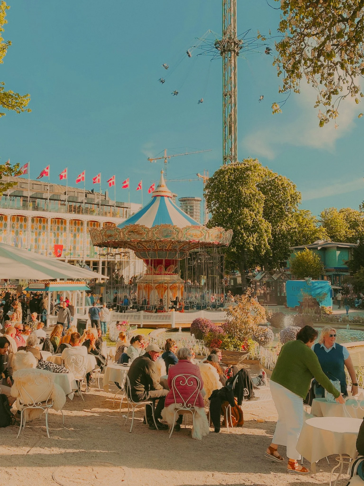 Une fête dans un parc d'attractions avec une grande roue, un carrousel, des gens assis à des tables, représentant une ambiance festive en plein air.