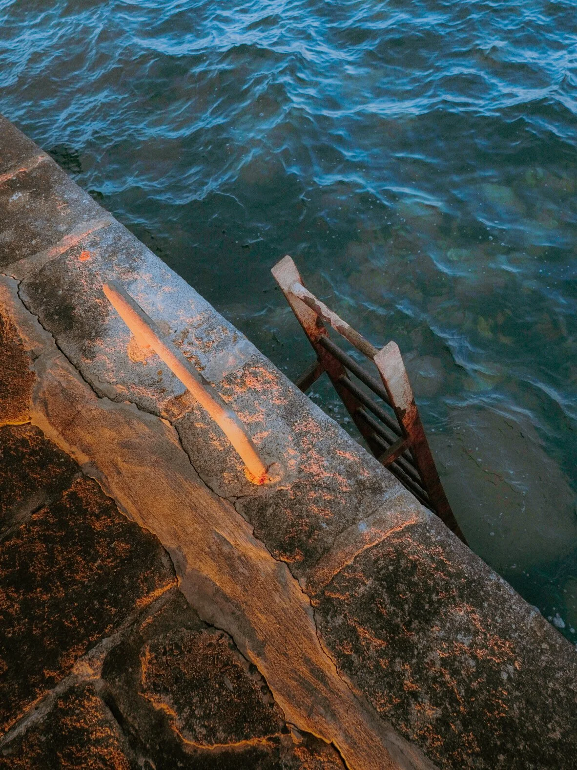 Escalier en métal rouillé menant dans l'eau, côté d'un quai en pierre avec des fissures, au coucher de soleil.