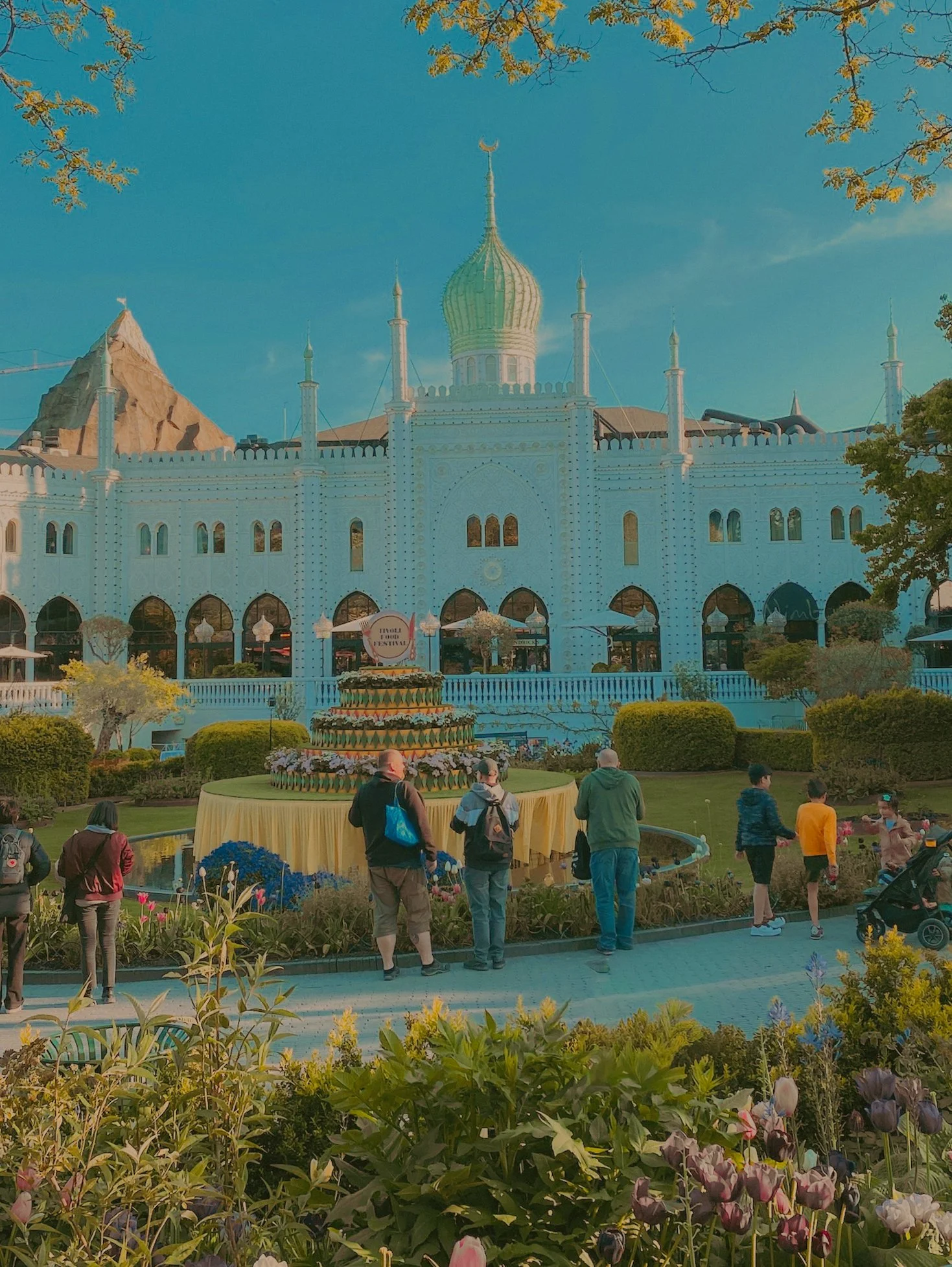 Un groupe de personnes devant un bâtiment blanc de style oriental, avec une coupole dorée et des minarets, entouré de jardins fleuris.