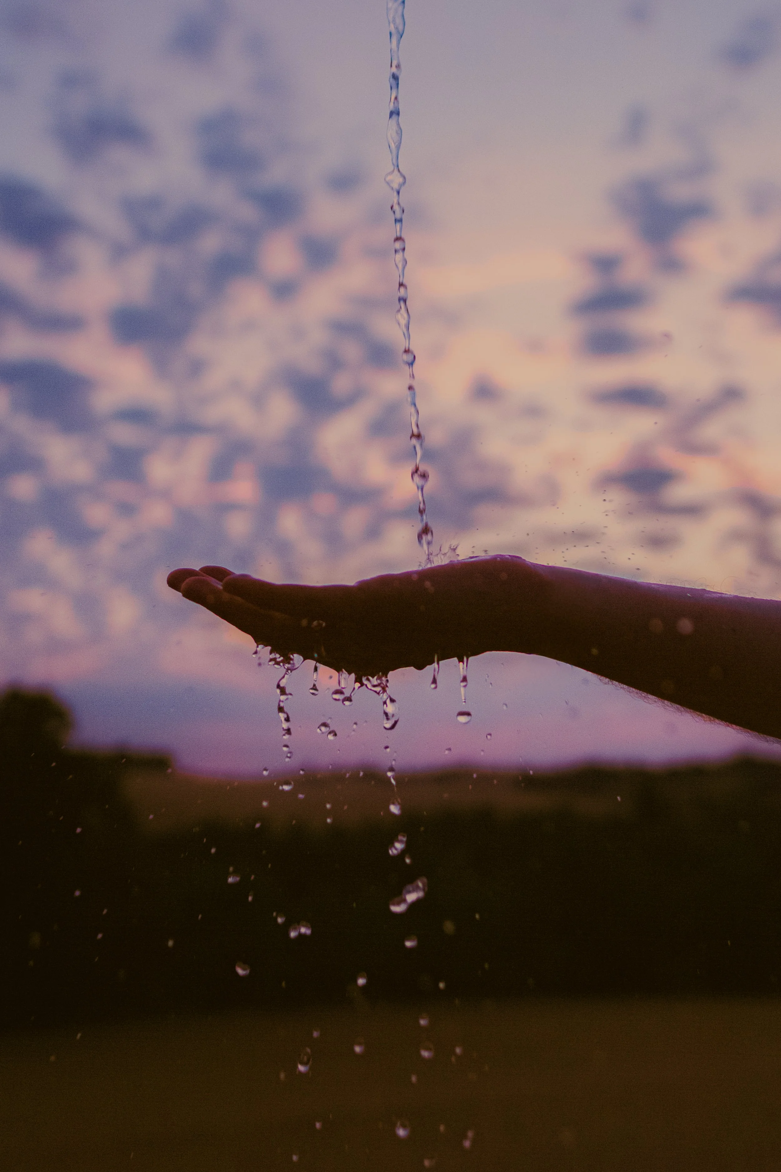 Une main ouverte sous une chute d'eau, avec un ciel coloré en arrière-plan au coucher du soleil.