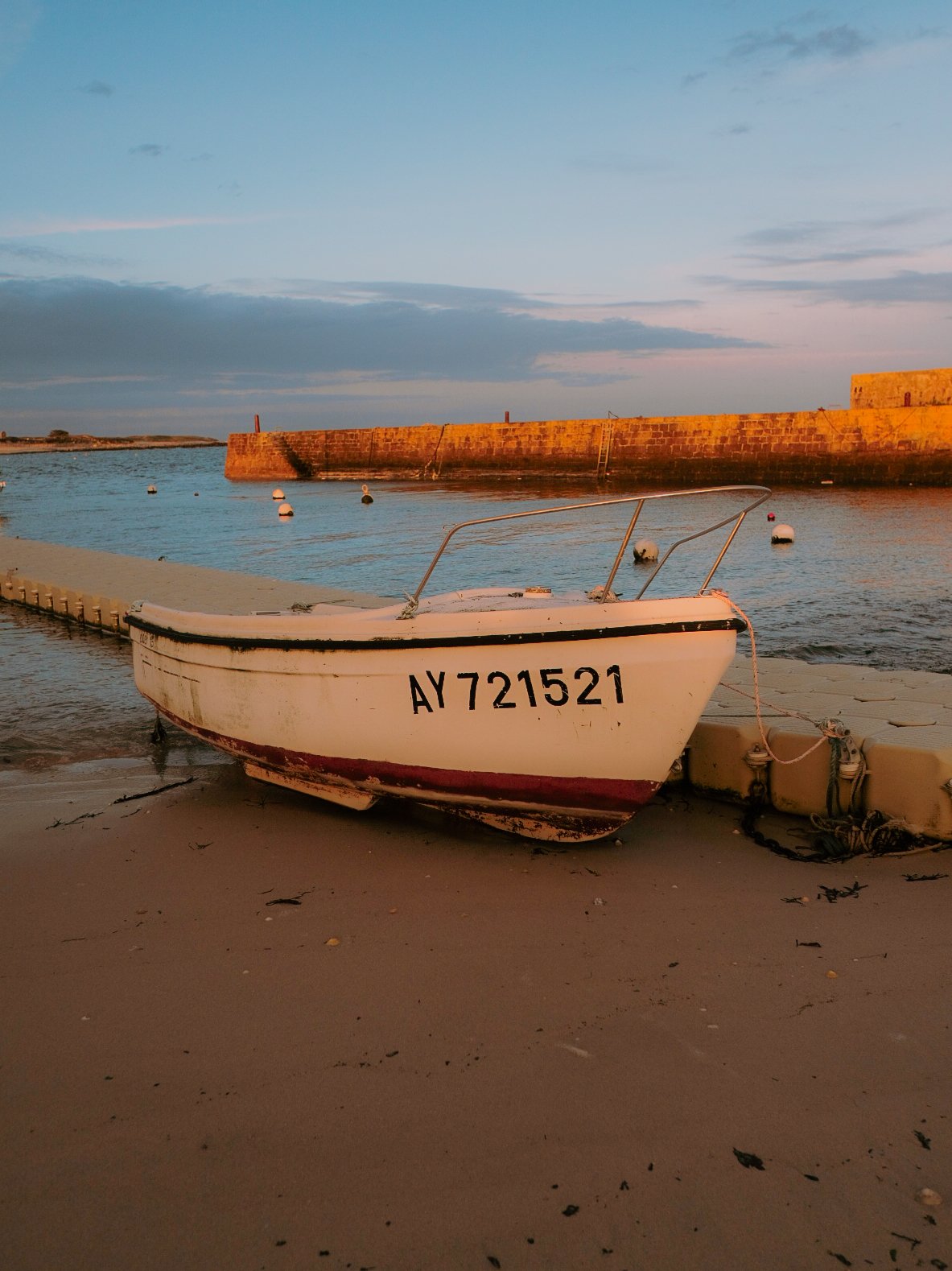 Petite bateau blanc avec l'inscription AY721521, échoué sur la plage au coucher du soleil, avec un quai en béton et un mur en pierre en arrière-plan.