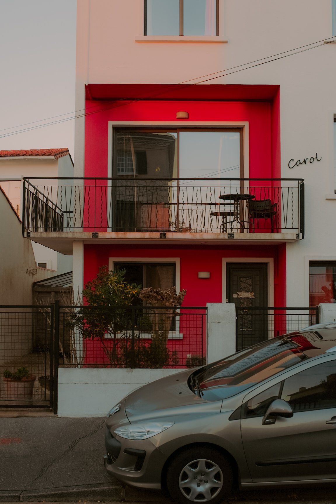 Un immeuble résidentiel à deux étages avec une façade rose, une petite terrasse au premier étage avec une table et des chaises, une voiture grise garée devant, et le nom « Carol » écrit sur le mur.
