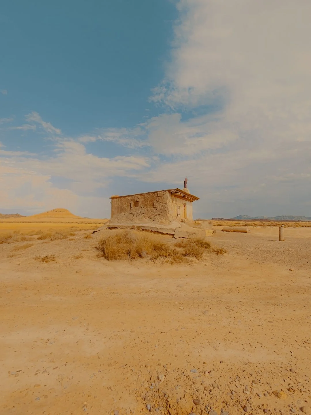 Petite maison en pierre dans un désert avec un ciel bleu et quelques nuages, entourée de buissons jaunes et de peu d'autres constructions.