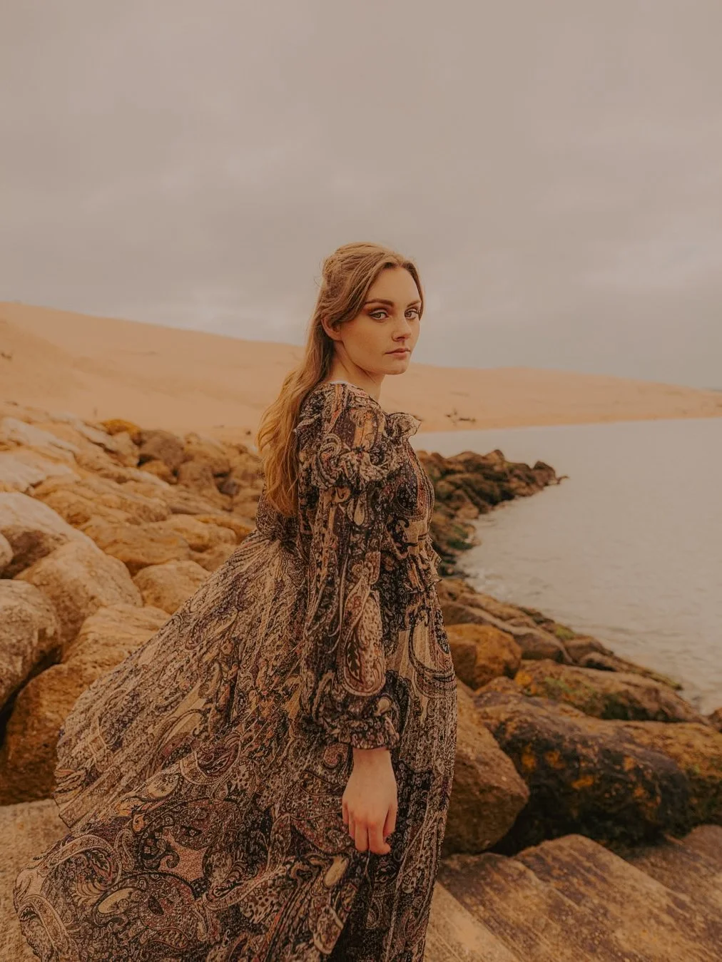 Une femme debout sur une plage rocheuse en pleine nature, portant une robe longue à motifs, avec un ciel grisâtre en arrière-plan.