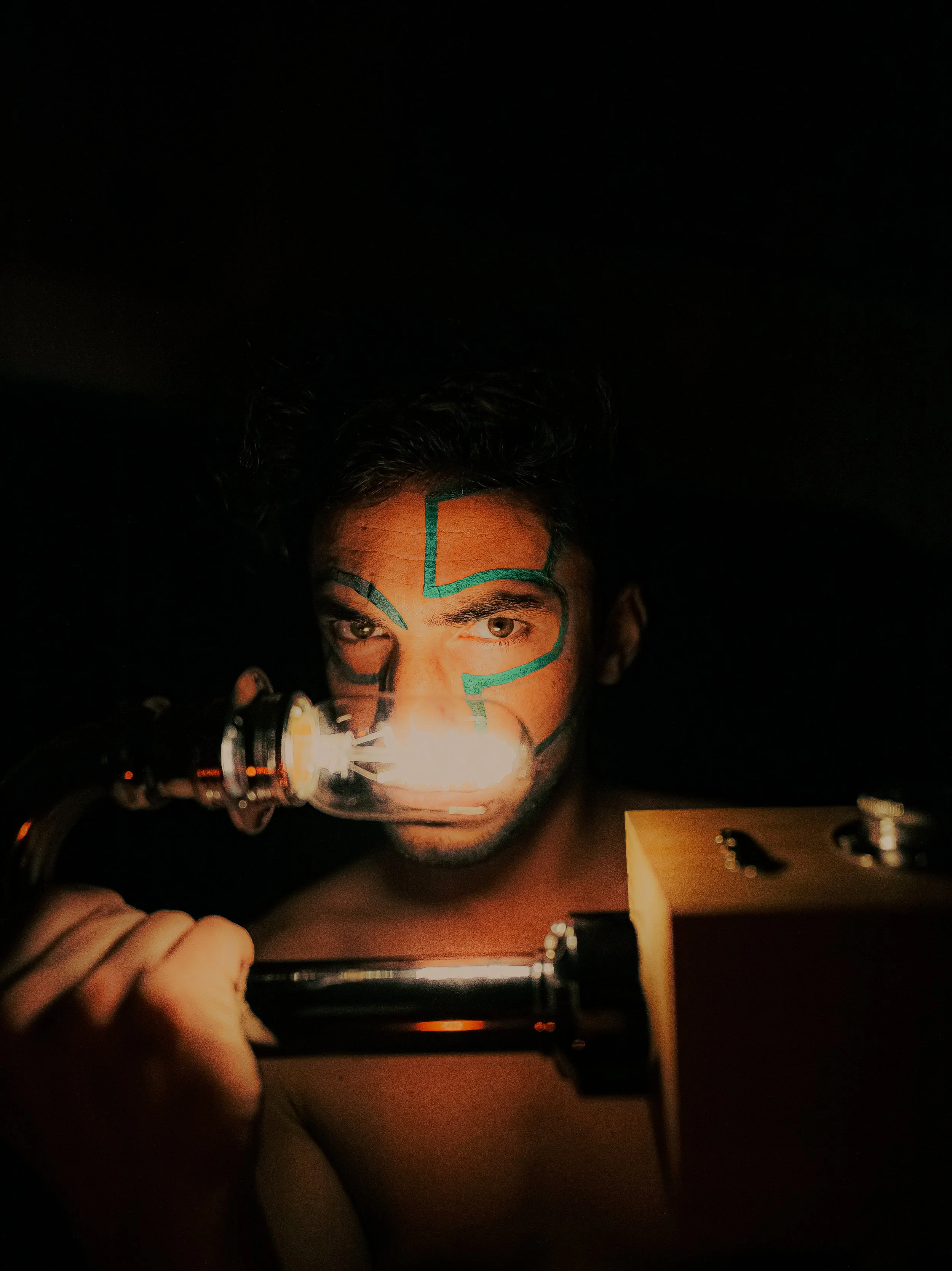 Jeune homme avec maquillage artistique face à une lampe à incandescence dans un environnement sombre.