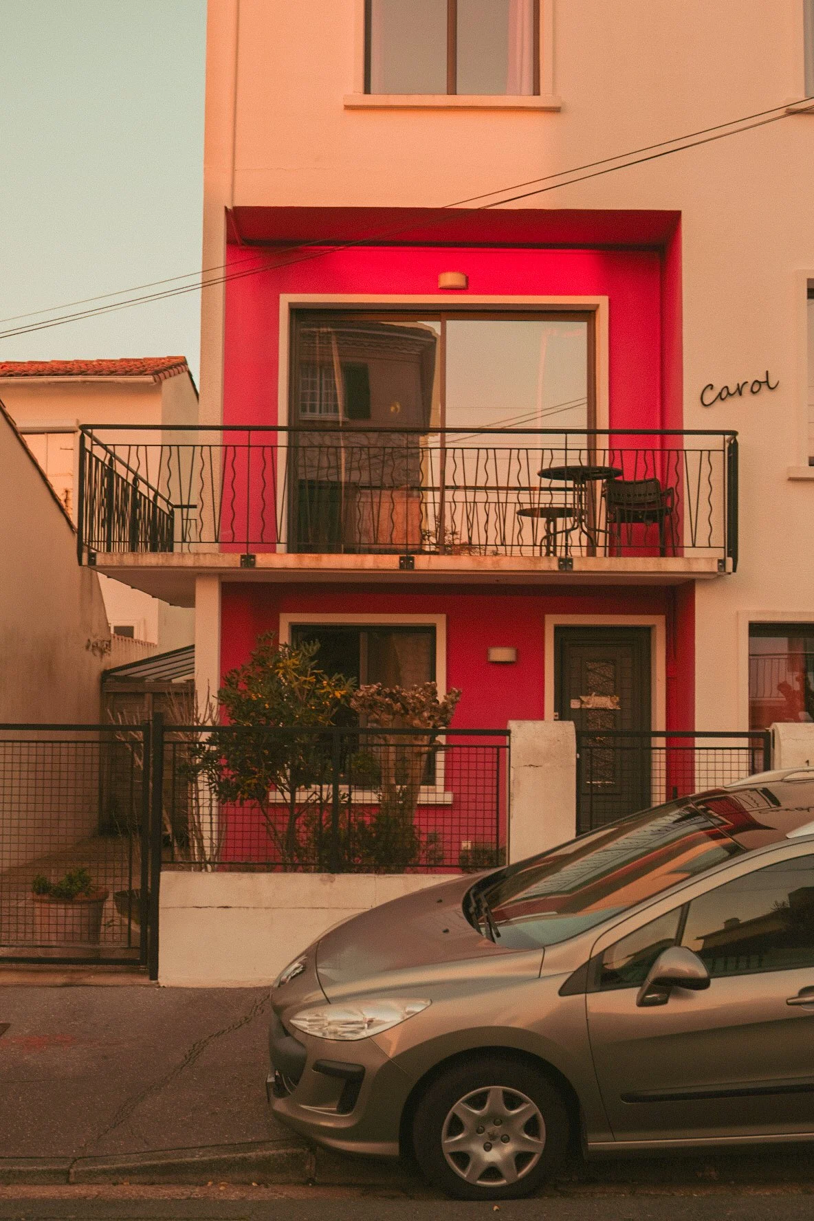 Un bâtiment résidentiel avec une façade rose et des balcons en fer forgé, un appartement au premier étage avec une table et des chaises sur le balcon, une voiture gris métal garée en premier plan, et le nom 'Carol' écrit sur la façade.
