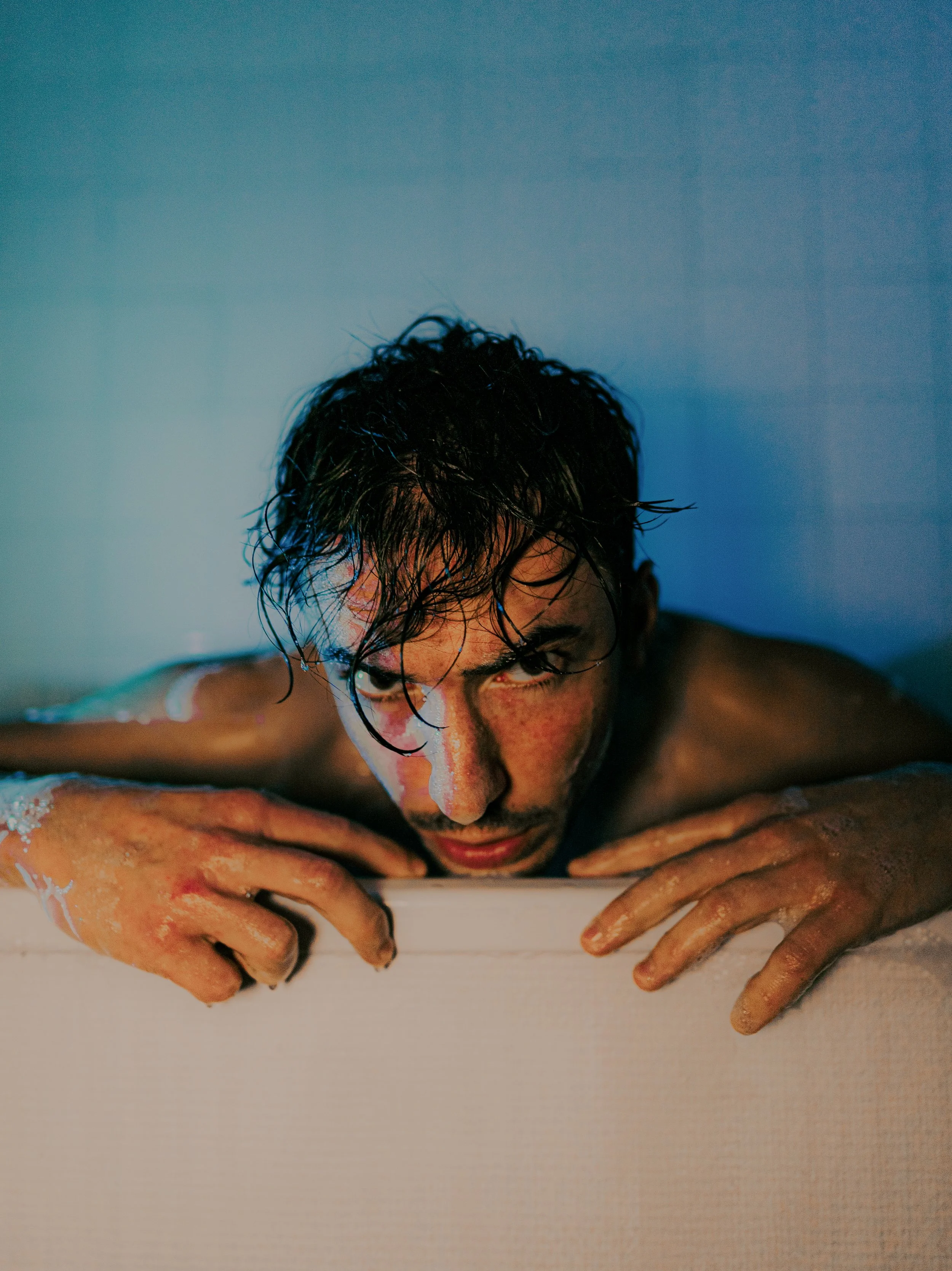 Un homme avec des cheveux bouclés, la tête reposant sur le bord d'une baignoire, regardant fixement l'objectif, avec de la mousse sur ses mains et son visage, dans un environnement de bain