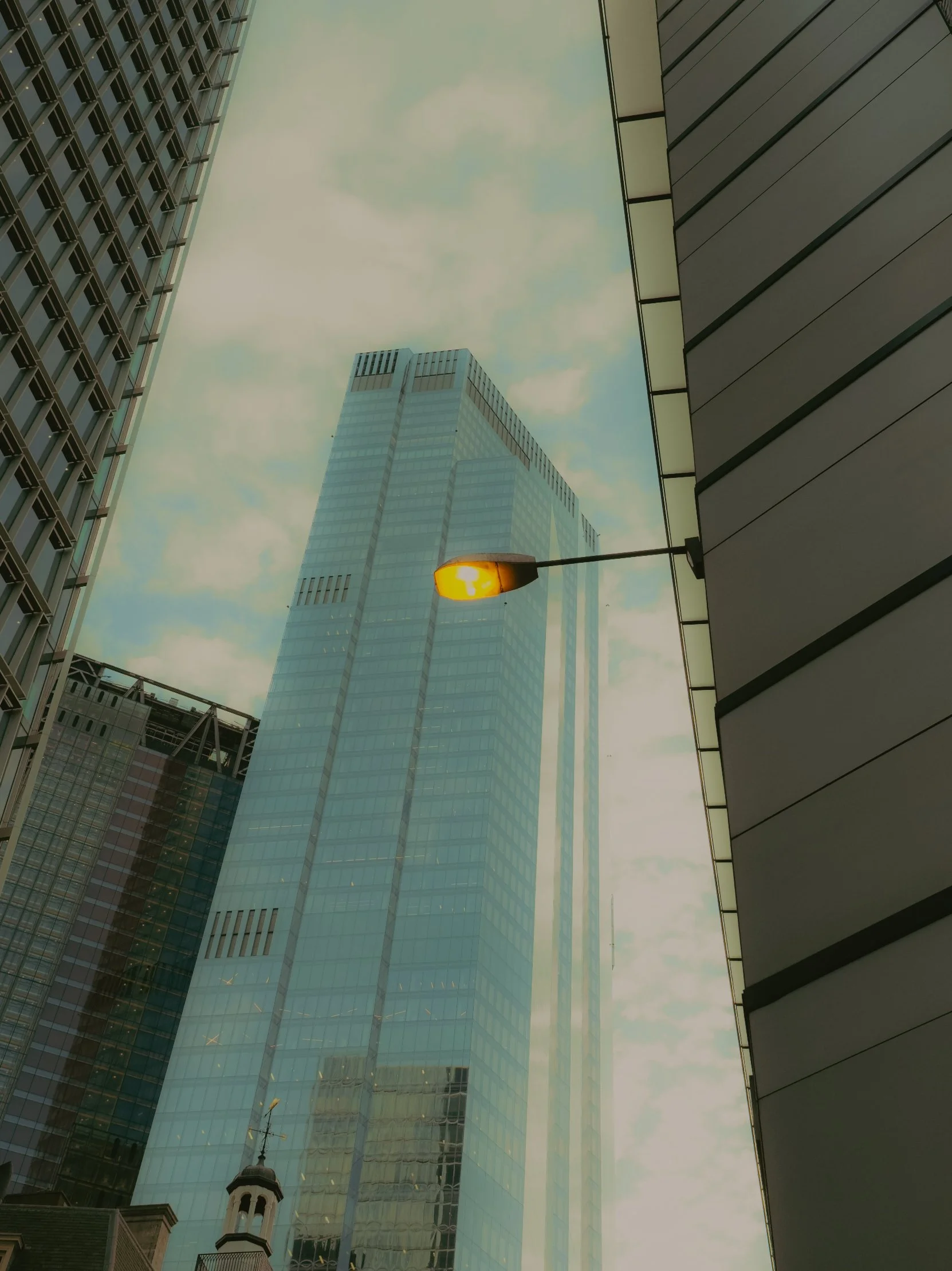 Vue du bas d'un gratte-ciel en verre dans une ville, avec un lampadaire orange en premier plan, entouré d'autres bâtiments modernes.