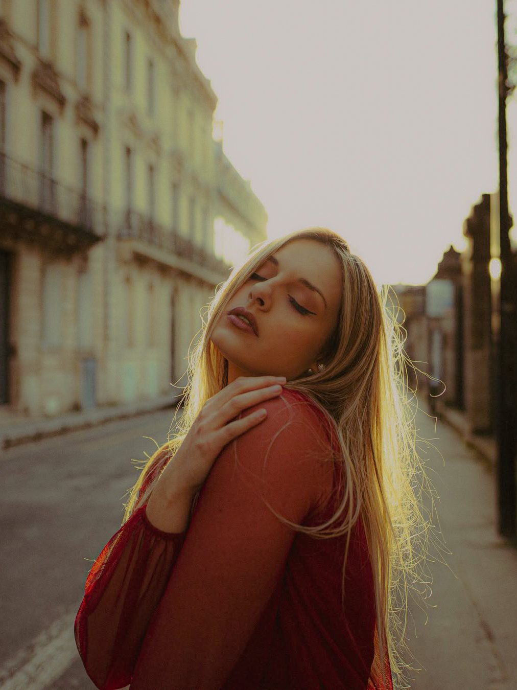 Une jeune femme avec des cheveux blonds, portant une robe rouge, se tient dans une rue pavée avec des bâtiments anciens en arrière-plan, lors du coucher du soleil.