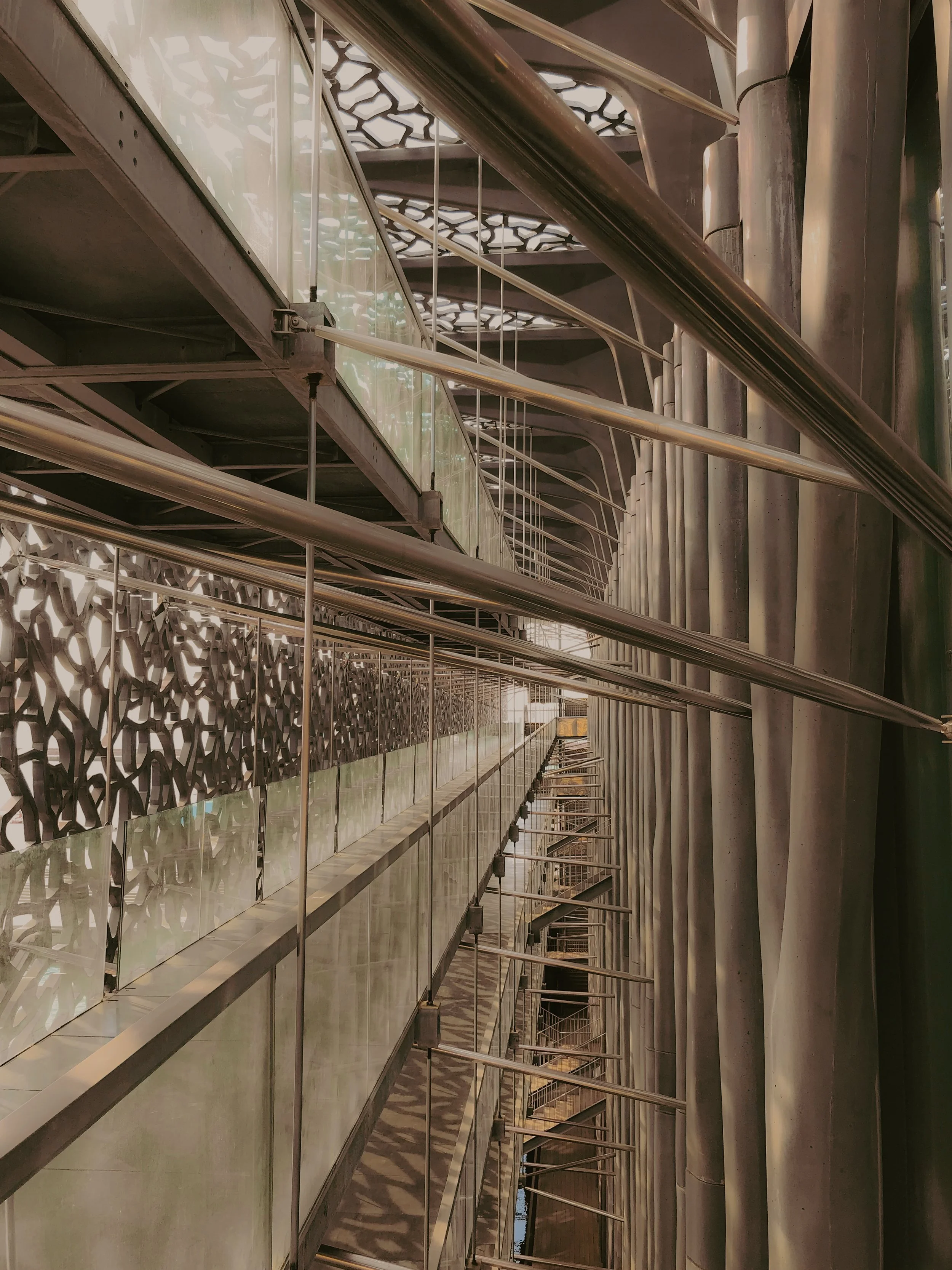 Vue de l'intérieur d'une structure architecturale moderne avec des rampes en acier, des murs en verre, et des éléments en métal décoratif, vue en perspective verticale.